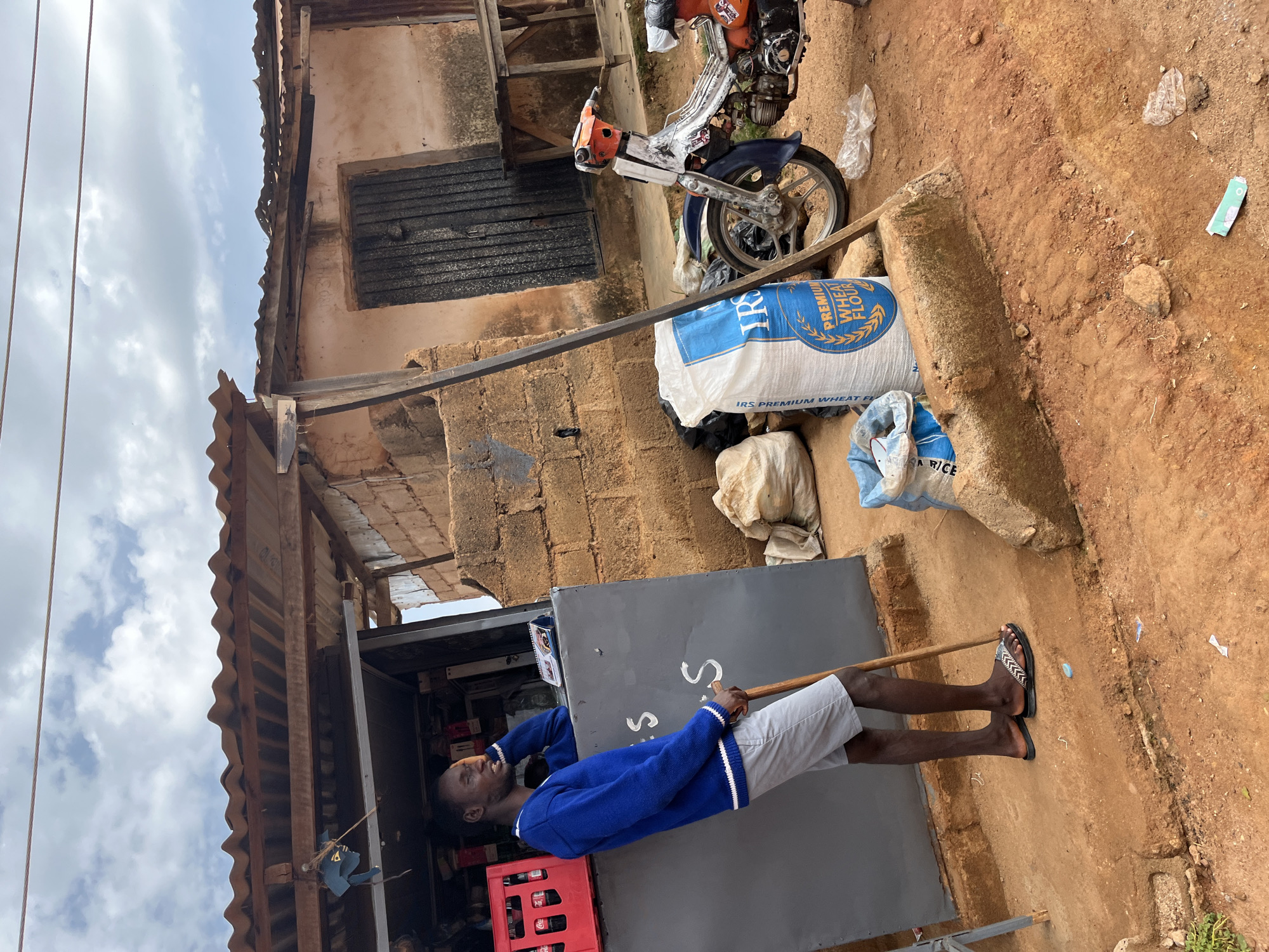 A young Black man, wearing a blue sweater and gray shorts, leans against a gray counter at a roadside stall. He holds a wooden stick and rests his head on his hand, looking to his left. Behind him, a red crate of Coca-Cola bottles is visible. To his right, several bags of goods, including a large white bag labeled "IRS PREMIUM WHEAT FLOUR", are stacked. A dirt path leads to the stall, and a red and silver motorcycle is parked nearby. The stall is constructed of brick and corrugated metal roofing. The scene appears to be a street in Jos, Nigeria, characterized by the unpaved ground and simple, open-air structures typical of local markets. The sky is partly cloudy.
