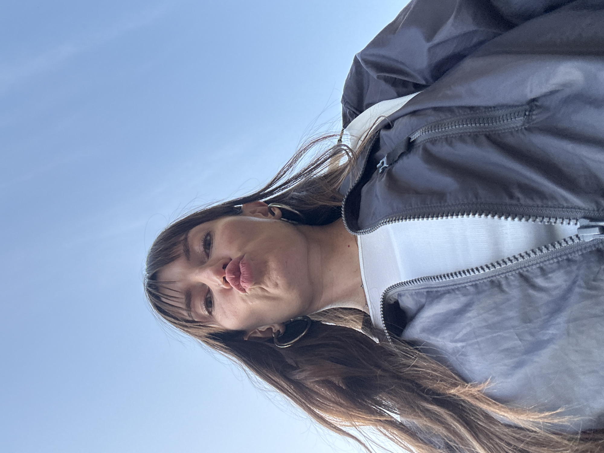 A low-angle, outdoor, close-up shot shows a young adult woman with long, dark brown hair, styled with bangs, wearing a white top and a dark grey zippered jacket. She is puckering her lips as if for a kiss, and her eyes are looking directly at the camera. She has large hoop earrings in both ears and a nose ring. The background is a clear blue sky. The image does not provide any specific details that would indicate it was taken in Moscow, Russia.