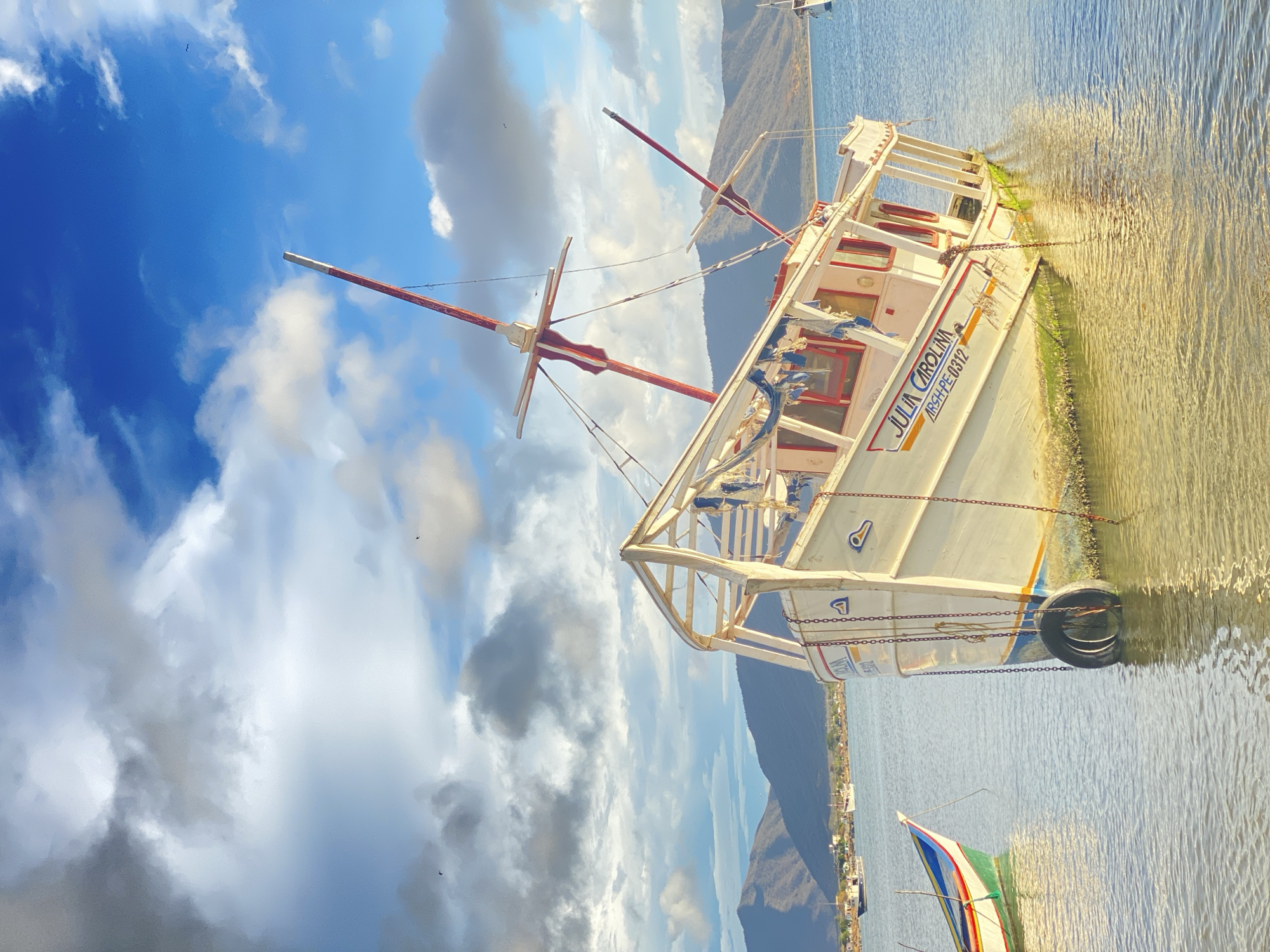 A white boat named "Julia Carolina" with red and yellow accents is docked on a body of water in Juan Griego, Venezuela. The boat has two red masts and a tire hanging from its side. There is a small building in the background, as well as a mountain. The sky is blue with white clouds. There is also another boat in the background.