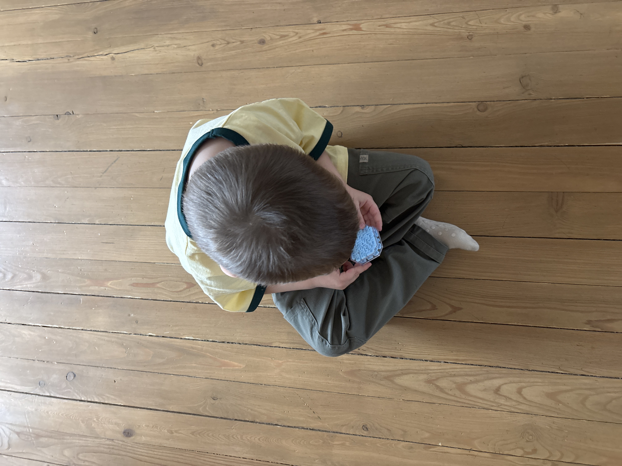 A high-angle shot depicts a child seated cross-legged on a wooden floor. The child wears a light yellow t-shirt with dark green trim and olive green pants, with a white sock visible on the right foot. The child is holding a small, light blue, possibly toy-like object, in their hands. The wooden floor is comprised of narrow planks and exhibits natural wood grain patterns. Based on visual data alone, it is not possible to establish that the location is Chaikovsky, Russia.