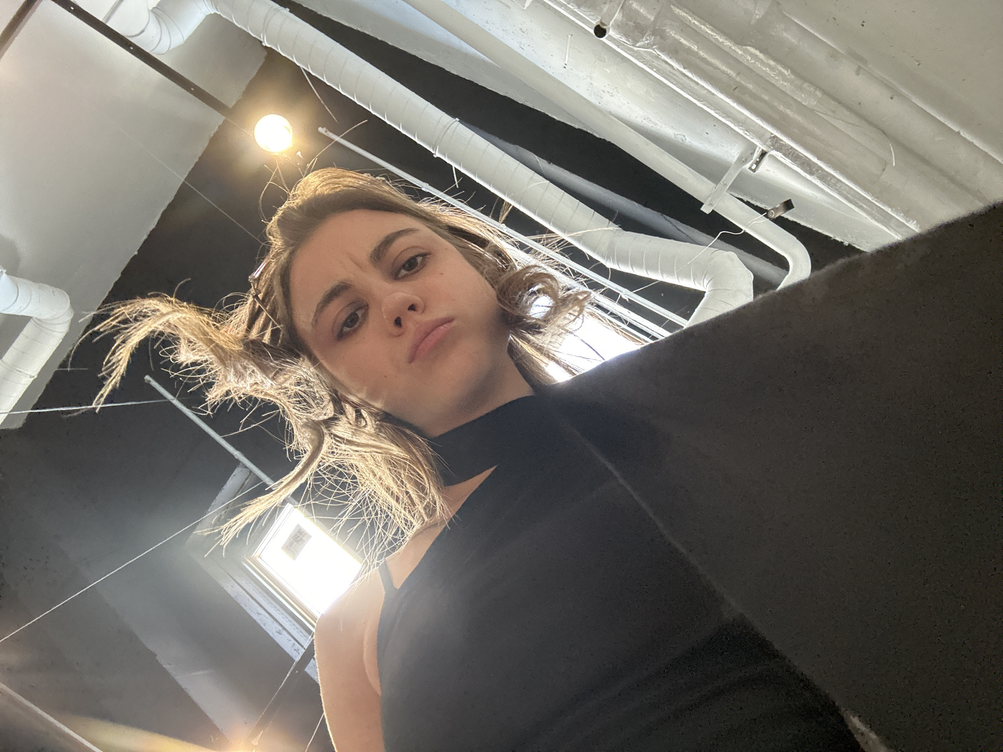 A low-angle shot shows a young woman with her hair blowing upwards. She is wearing a black top and looking down at the camera with a neutral expression. The background consists of industrial piping and a ceiling with exposed light fixtures, suggesting an indoor urban environment. The lighting is somewhat dim with a spotlight effect from above. The image appears to have been taken in Moscow, Russia.