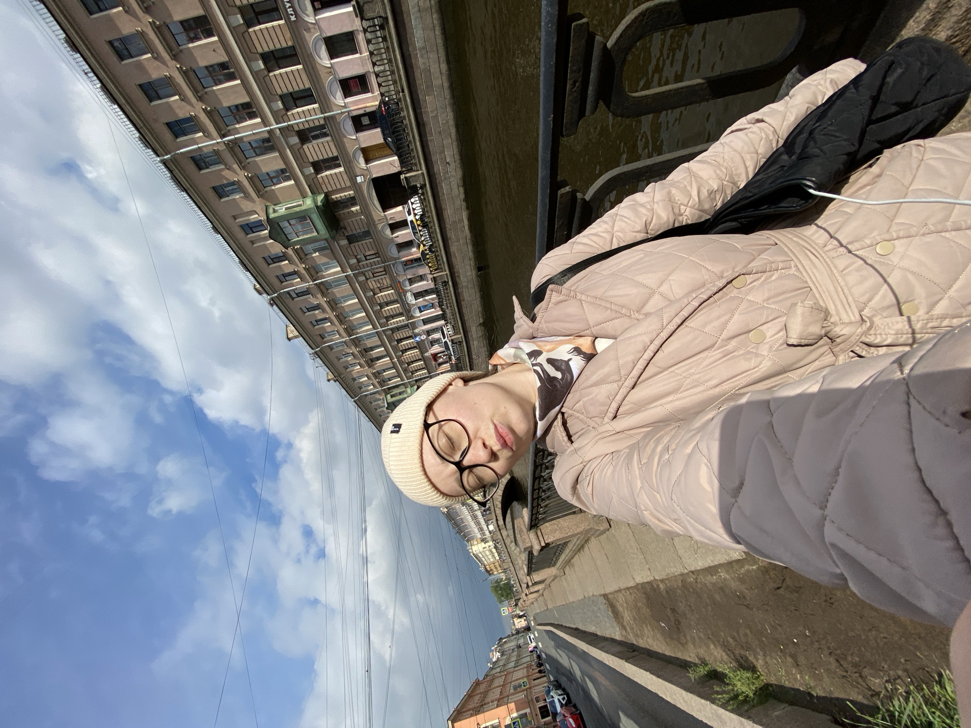 A rotated eye-level shot shows a person in a beige quilted coat and white beanie with round glasses, posing along a bridge in Saint Petersburg, Russia. The bridge's wrought-iron railing is on the right. Behind the railing is the Moika River. On the left side of the bridge is a street with buildings and cars. In the background is a sky with clouds. The architectural style and colors of the buildings are typical of Saint Petersburg.
