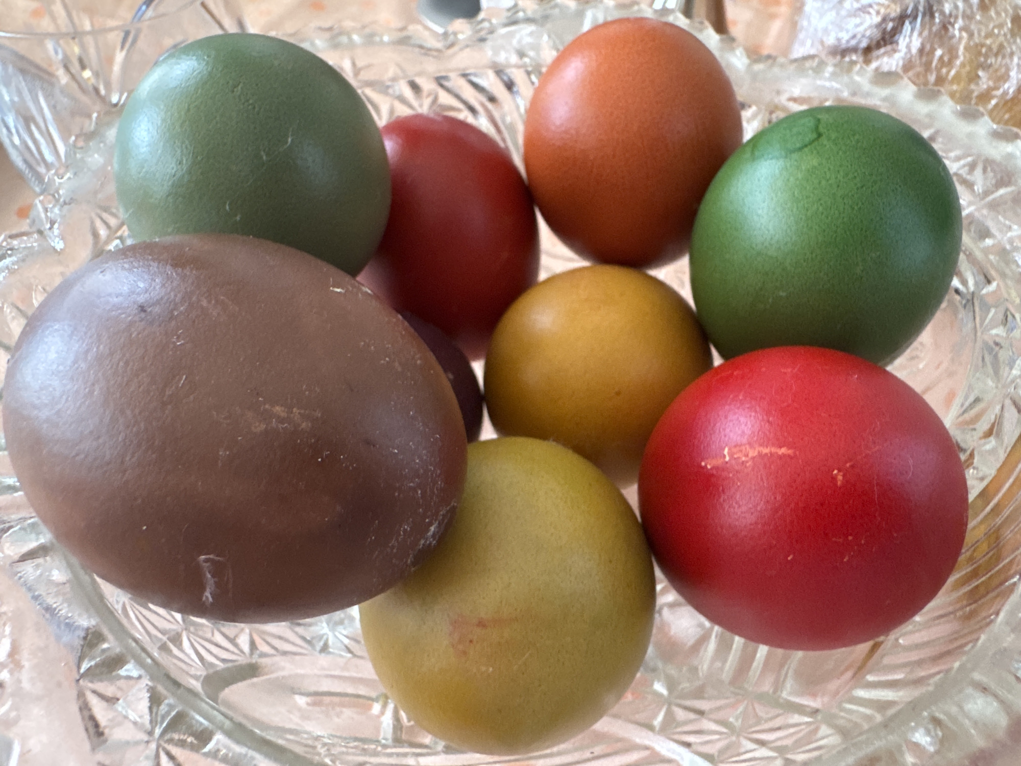 The image shows a close-up of nine colorful, dyed Easter eggs in a clear, cut-glass bowl. The eggs are arranged in a pile, with colors including brown, red, orange, yellow, and green. The bowl sits on a surface with a blurred background. There are no people visible in the image. The scene suggests a domestic setting, likely during or after Easter celebrations. There are no specific visual cues that connect this image to Sofia, Bulgaria.