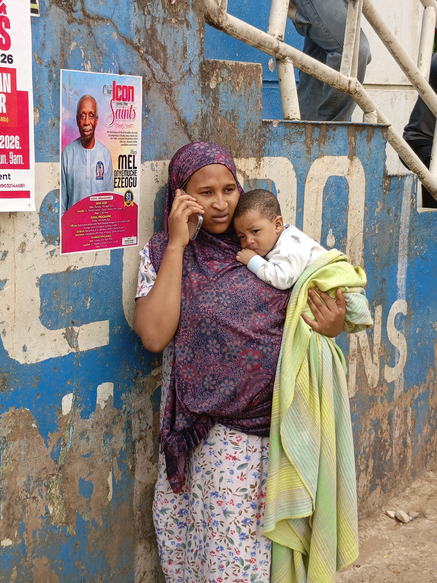 A woman stands against a blue wall with white lettering. She wears a floral dress and a patterned headscarf, holding a baby wrapped in a striped blanket. She is on a phone call. Behind her are two posters. One features a man's portrait with the text "Our Icon joins the Saints," including details about Mel Onyemuche Ezeogu's burial. The other poster shows "S" "26" "IN" and an unreadable message. Above the woman and the wall is part of a metal railing structure with a person in jeans visible in the background. The location is likely a street in Unknown, Unknown.