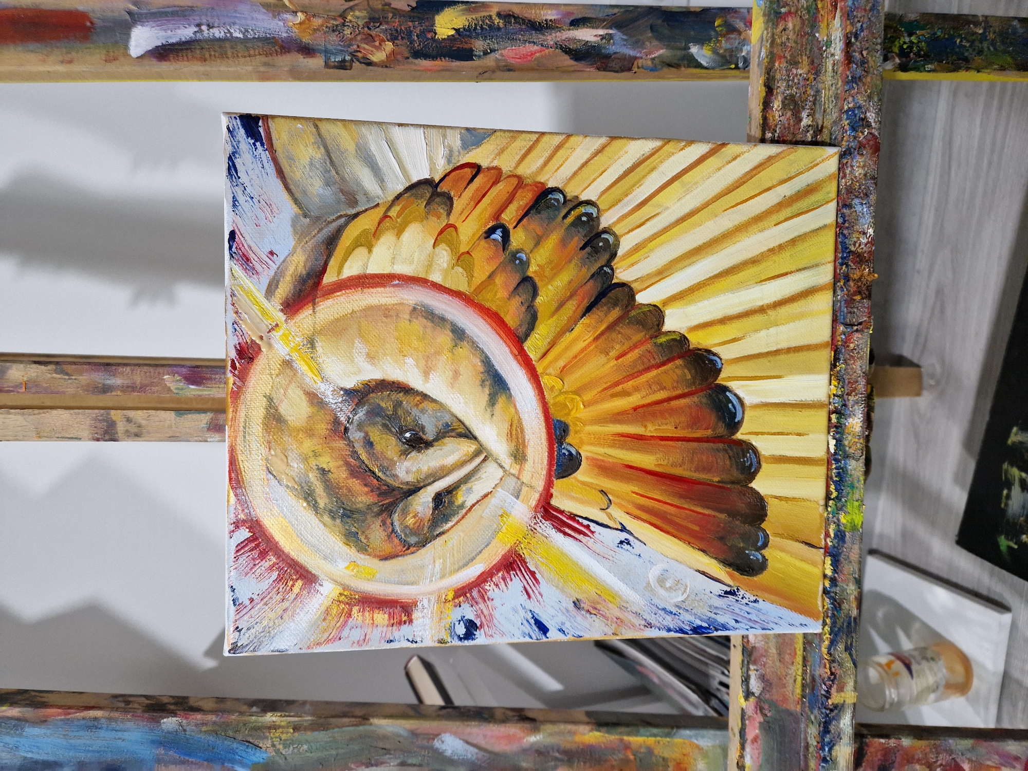 A square canvas painting is displayed on an easel. The painting features a stylized depiction of a sunflower seed head with golden and brown hues. A central circular form contains curled, dark shapes, resembling the seeds. The outer area of the seed head is surrounded by petals radiating outwards in shades of yellow, orange, and brown, with some ending in black tips. The background has areas of white and blue paint, providing contrast. The easel itself is splattered with various colors of paint, adding a textural element to the scene. The location is Unknown, Unknown.