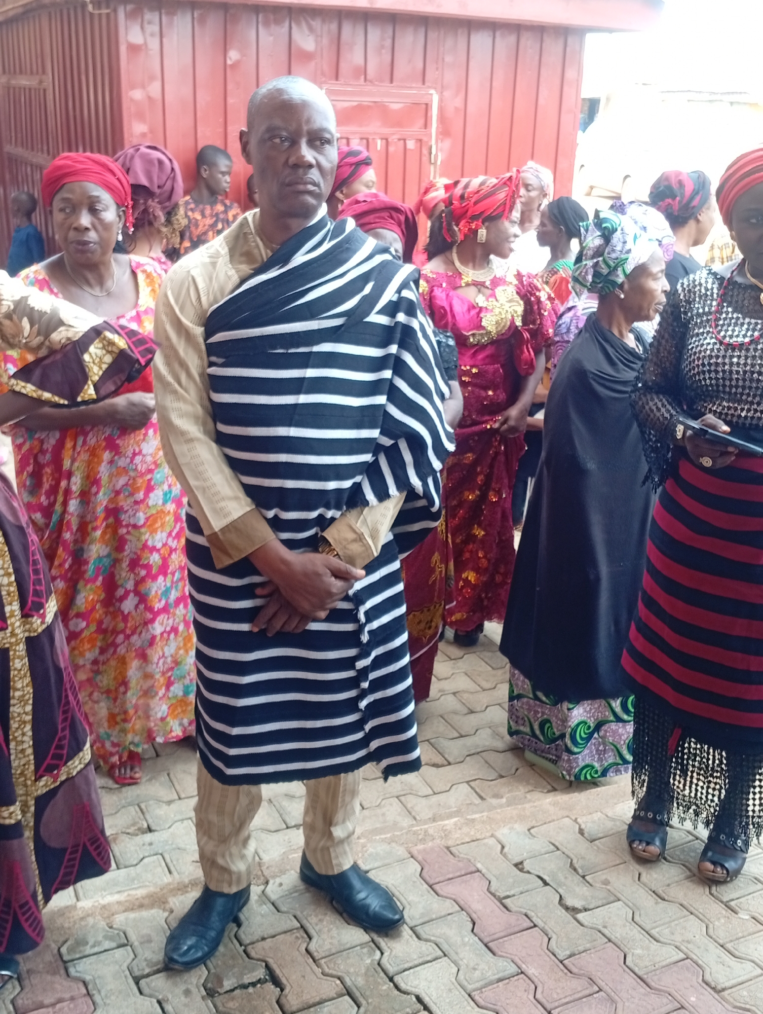 The image shows a group of people standing outdoors on a paved area, likely a gathering or event.
A man stands prominently in the foreground, wearing a light-colored shirt and trousers with a black and white striped cloth draped over his shoulders. He has dark blue dress shoes. Several women stand behind him, dressed in vibrant traditional clothing, including patterned dresses and headwraps.
The location appears to be an urban area, with a building in the background painted in a reddish-brown color. The paved area suggests a sidewalk or public space.