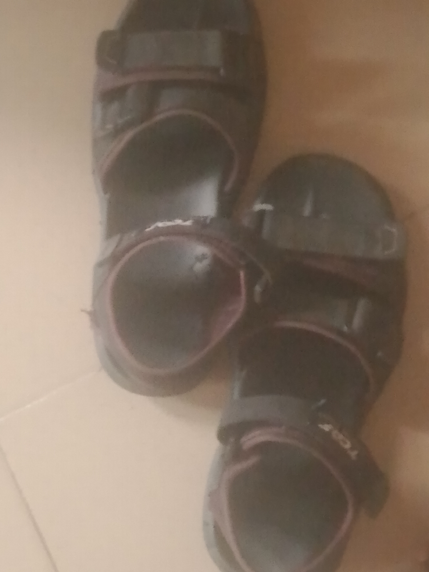 The image shows a pair of dark-colored sandals with red trim. The sandals appear to be made of synthetic materials, with adjustable straps. They are positioned on a light-colored floor, possibly tile or linoleum, with a hint of what looks like cardboard or a similar material in the background. The lighting is dim, casting shadows that obscure some details. The overall scene is unremarkable and lacks any specific indicators to place it in Jauro Yinu, Nigeria or any other particular location. There are no people visible in the image.