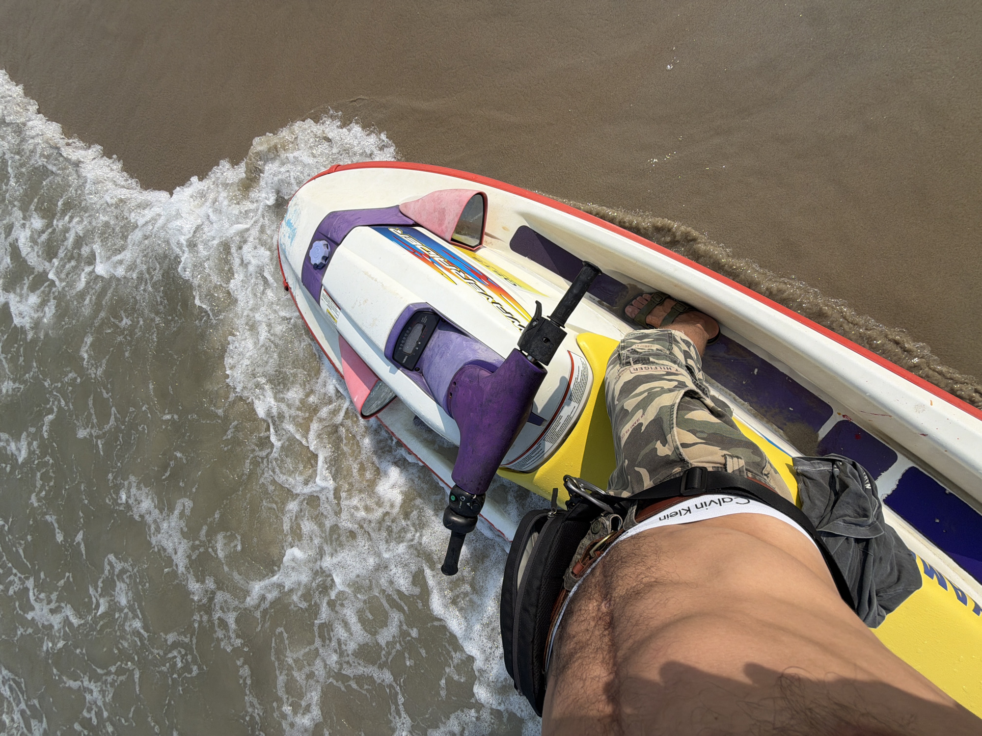 A high-angle shot shows a person on a white, red, and yellow WaveRunner-style personal watercraft in Đà Nẵng, Vietnam. The craft is partially submerged in foamy seawater near a beach. The person wears camouflage pants and brown sandals and is also wearing visible Calvin Klein underwear. A black bag is attached to the person's side. The water transitions from foamy waves to sandy shoreline in the background.