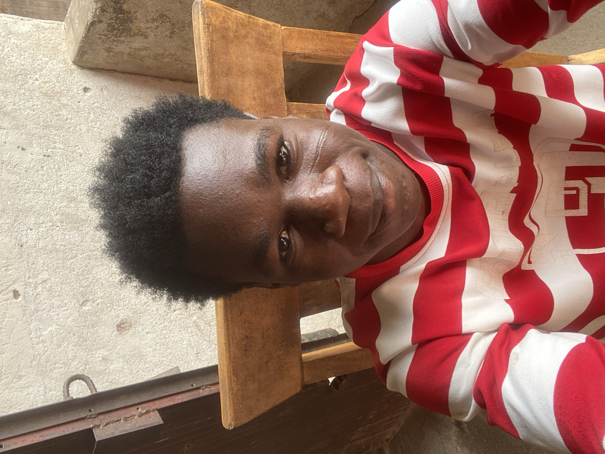 A medium shot of a dark-skinned person, likely male, with short curly hair, taken indoors. The person is wearing a red and white striped sweater, possibly seated on a wooden chair. The background shows a concrete wall and what appears to be an older, perhaps weathered, structure. The location is assumed to be Mararaba, Nigeria. The person is looking directly at the camera with a slight smile.