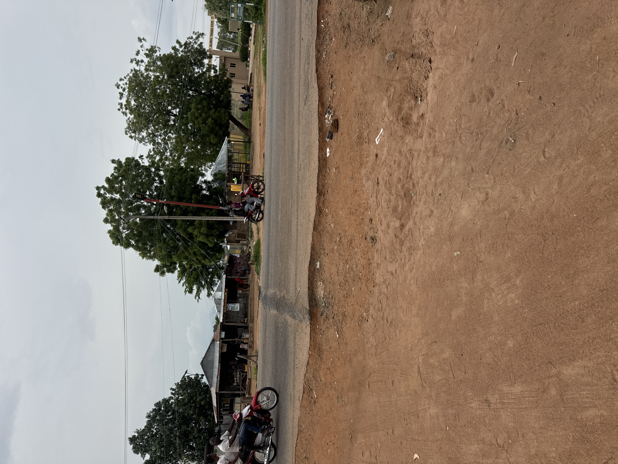 In Jauro Yinu, Nigeria, a red motorcycle with two male occupants rides on a paved road. The rider wears a white t-shirt and blue jeans, while the passenger wears a white t-shirt. To the left of the road, a reddish-brown dirt embankment slopes down. Across the road, a row of small, simple structures with corrugated metal roofs are visible, appearing to be shops or market stalls. Beyond these structures, several large green trees stand. In the background, a sign reads "FEDERAL POLYTECHNIC JALINGO, TARABA STATE". Several people are seated outdoors near a building in the distance, and more trees line the horizon under an overcast sky. Debris, including plastic bags and a rock, is scattered on the dirt embankment in the foreground.