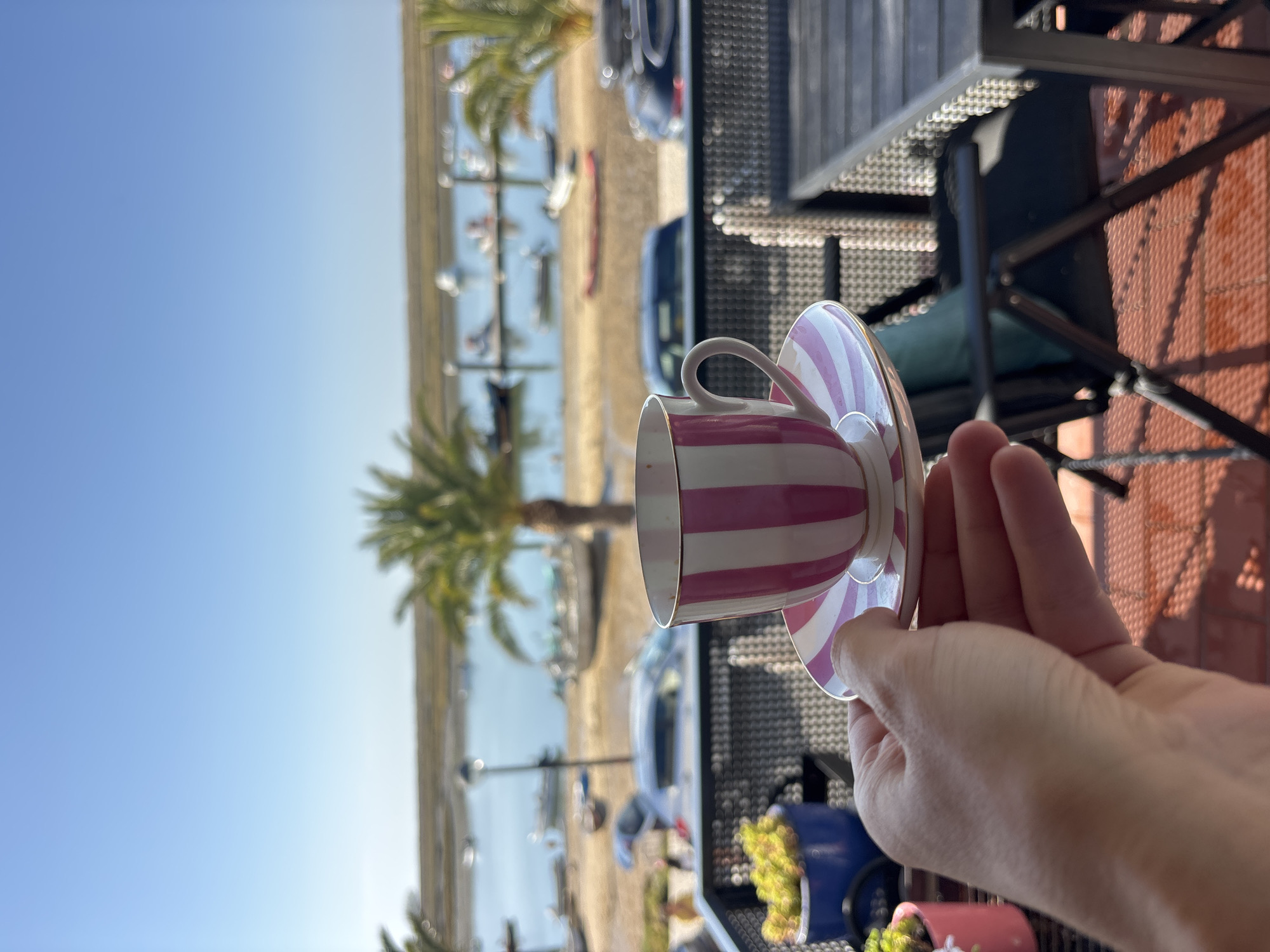 A hand holds a pink and white striped teacup and saucer at a balcony overlooking a coastal town. In the background, boats are moored in calm water near a sandy shore with palm trees and buildings. Cars are parked along the waterfront. A table and chairs are visible on the balcony. The sky is clear and blue. The location is identified as Tavira, Portugal.