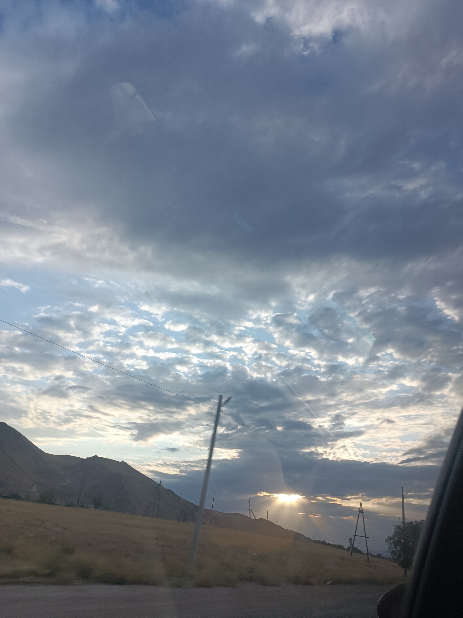The image captures a landscape scene near Sasyk-Bulak, Kyrgyzstan, dominated by a dramatic sky filled with textured clouds. Sunlight streams through gaps in the clouds, creating visible rays that illuminate parts of the sky and landscape. In the foreground, a dry, grassy hillside slopes upwards, with several utility poles scattered across it. The terrain appears arid, with muted brown and yellow tones. In the background, a mountain range rises, its slopes appearing barren and sparsely vegetated. A road is visible in the very bottom of the frame. The overall atmosphere is one of natural beauty and potentially a transition in weather, given the interplay of sun and clouds.