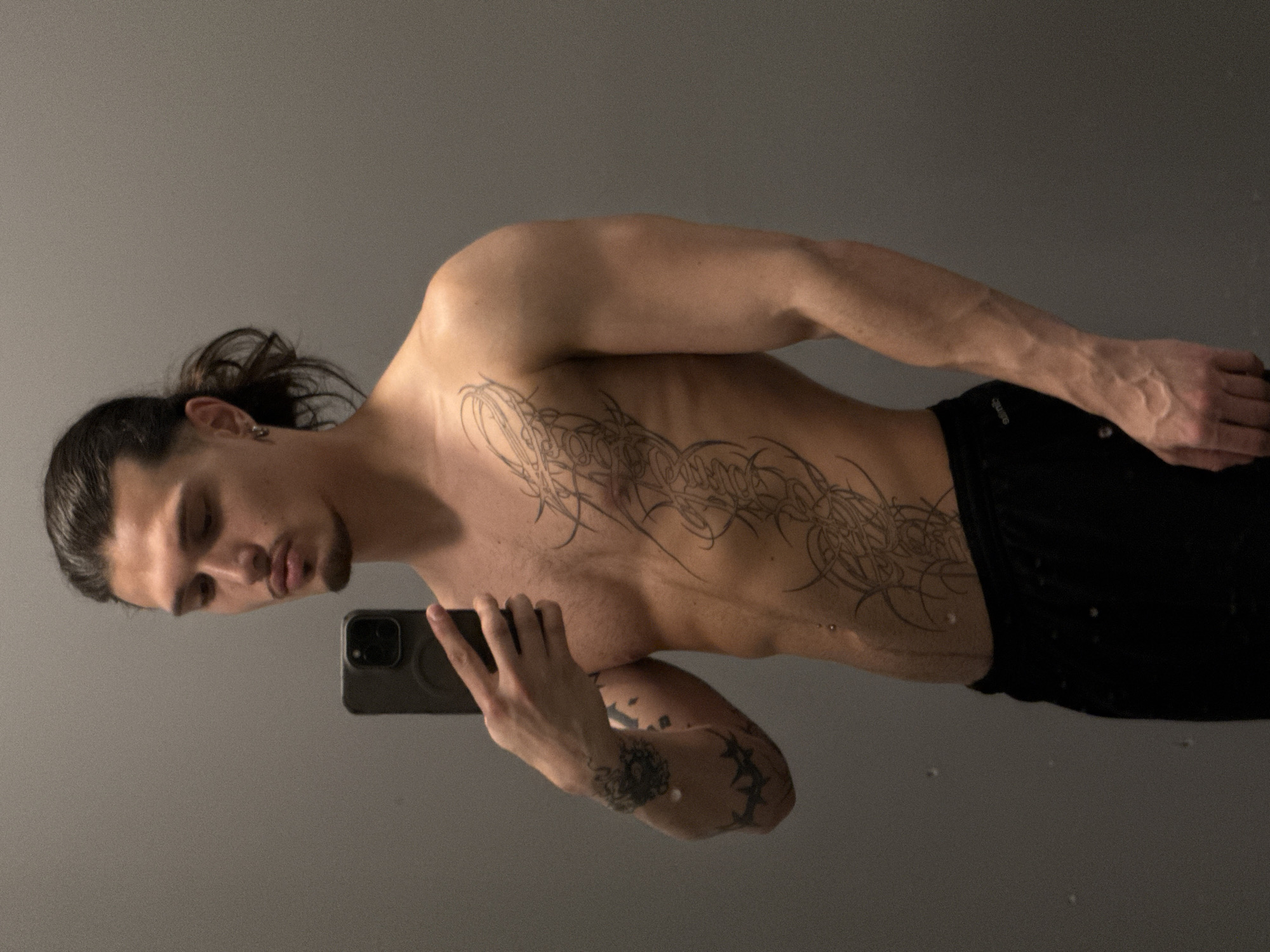 The image shows a young man with dark hair pulled back in a ponytail, taking a selfie in a gym locker room. He is shirtless, revealing a tribal tattoo that extends from his chest down his abdomen, and tattoos on his left arm. He is wearing black athletic shorts. The background is a plain gray wall, typical of a gym environment. The lighting is overhead, casting soft shadows. No specific location in Sofia, Bulgaria is identifiable.