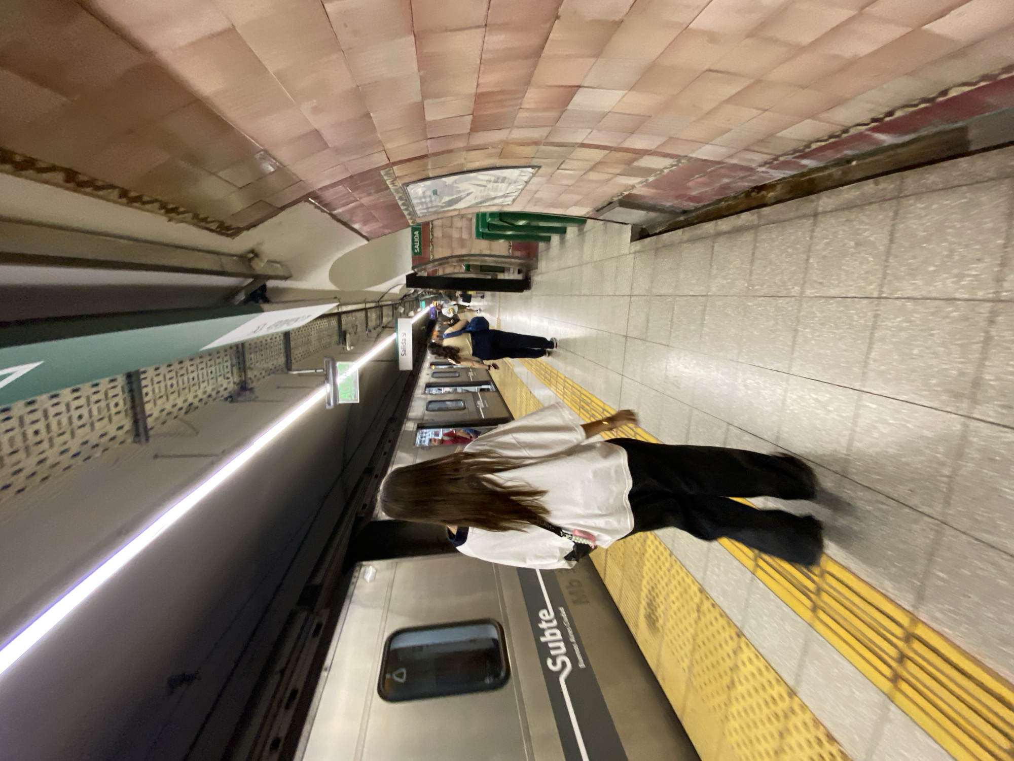 The image captures a scene inside a subway station in Buenos Aires, Argentina. The arched ceiling above is tiled in light brown and beige hues. On the right side of the frame, a person is standing on the platform, their back towards the camera. They are wearing a white top, black pants, and have long brown hair. The platform edge is marked with a yellow textured strip. To the left, a subway train is present, its silver exterior bearing the word "Subte" and additional text below. Further in the distance, two individuals appear to be lying down on the platform. Signage with the word "Salida" is visible in the background.