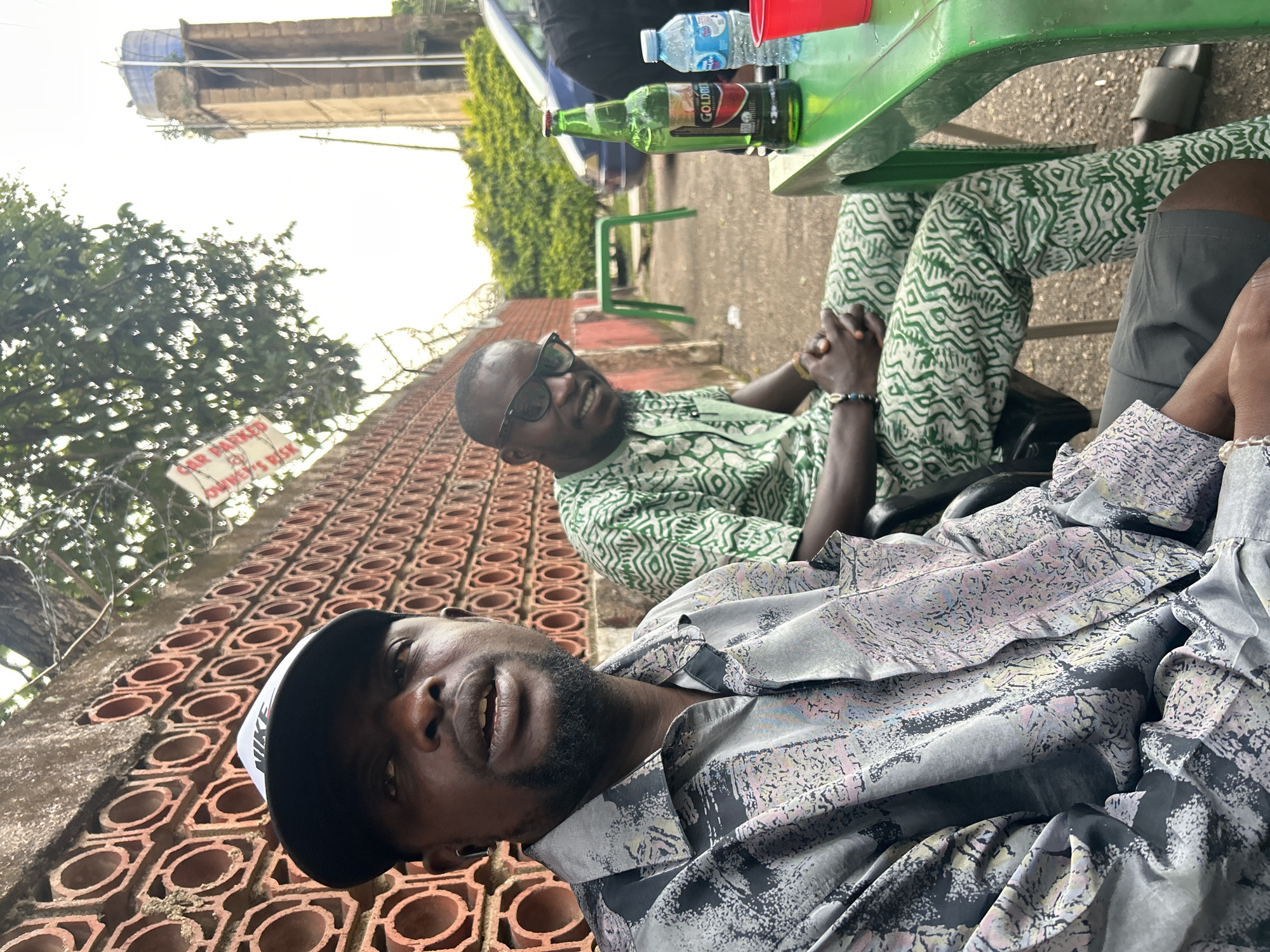 In Jos, Nigeria, two men are seated at a green plastic table outdoors, situated in front of a red brick wall with a patterned design. The man in the foreground, on the left, wears a grey patterned shirt and a black Nike baseball cap. He has a beard and is looking slightly up and to his left. To his right, the second man is dressed in a green and white patterned outfit and wears black-rimmed glasses. He is looking towards the viewer and smiling. On the table between them sits a green bottle of Goldberg beer and a bottle of water, alongside a red plastic cup. In the background, a tall, weathered concrete tower with a blue water tank on top is visible, along with a blue car and dense green foliage. A white sign with red lettering, partially obscured by leaves, is attached to a barbed wire fence to the left of the frame. The overall setting suggests a casual outdoor gathering.