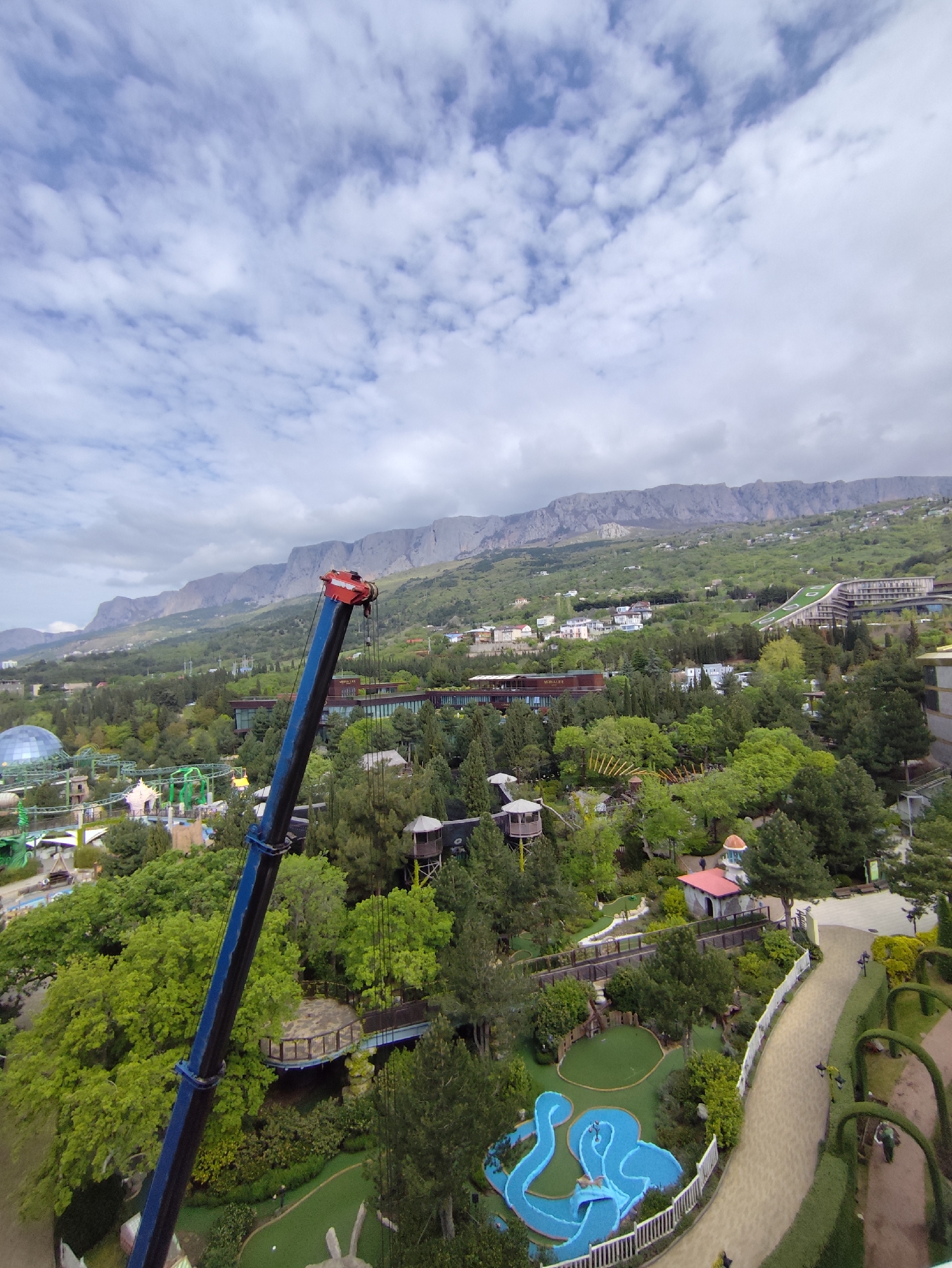 A high-angle, wide shot depicts a landscape in Симеизский поселковый совет, Ukraine under a partly cloudy sky. A mountain range stretches across the upper portion of the image, serving as a backdrop.
The foreground is dominated by lush greenery with trees and gardens. A blue crane boom stretches diagonally across the left side of the frame.
In the middle ground, various structures including buildings and attractions of a park are nestled amongst the trees. A mini-golf course with blue water features and white fences is visible. Walkways and paths meander through the green spaces.