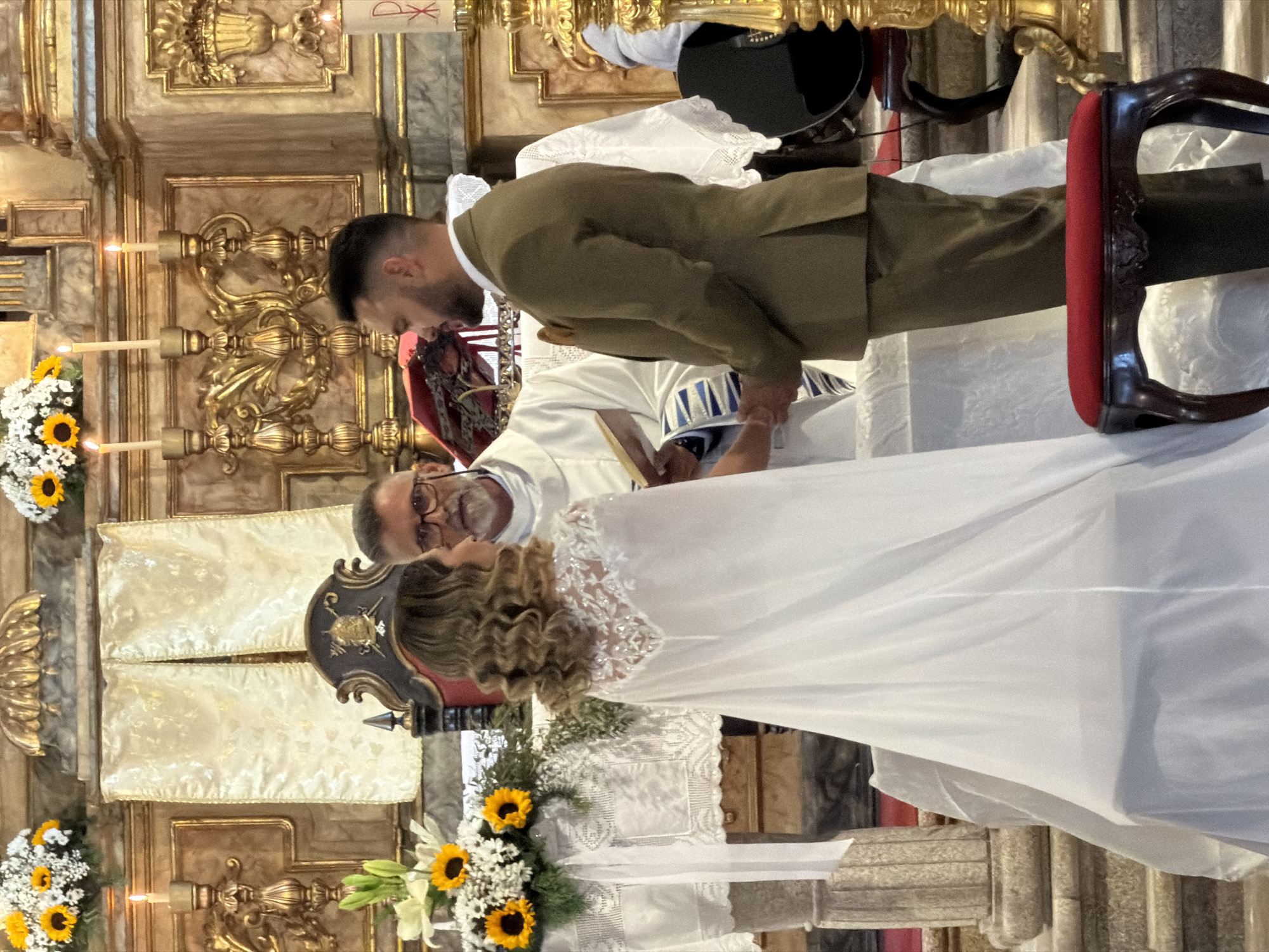 A wedding ceremony is taking place in a church. A bride in a white gown and a groom in a green suit are holding hands in front of a priest who is reading from a book. The church has a gilded altar and is decorated with sunflowers and white lilies. Candles are lit on the altar. The image was likely taken in a church in Vila Real, Portugal.
