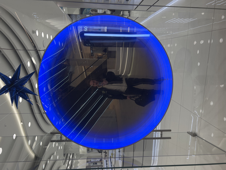 The image shows a person's reflection in a circular, blue-lit structure, likely an art installation or design element. The reflection shows the person taking a picture and includes details of the building's interior. A blue star-shaped decoration hangs nearby, while the surrounding environment is a modern, well-lit shopping mall. The circular structure is suspended, offering a unique perspective of the mall's architecture.