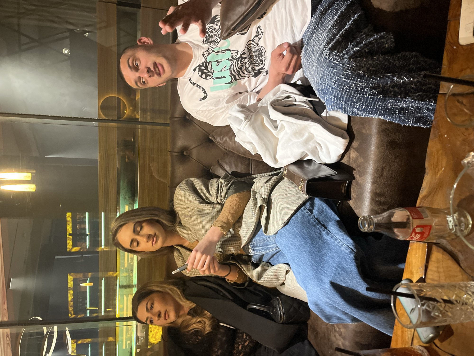 The image depicts a group of three people seated on a brown leather couch in an indoor setting, possibly a bar or lounge, in Burgas, Bulgaria. A man in a white t-shirt with tiger graphics is seated at the top, gesturing with his right hand. To his right is a blue textured jacket. Below him, a woman in a plaid blazer is holding a vape pen. Another woman wearing a black top and jacket sits next to her. A table to the right displays an empty Coca-Cola bottle and glasses with straws. A window or glass panel reflects the interior of the establishment.