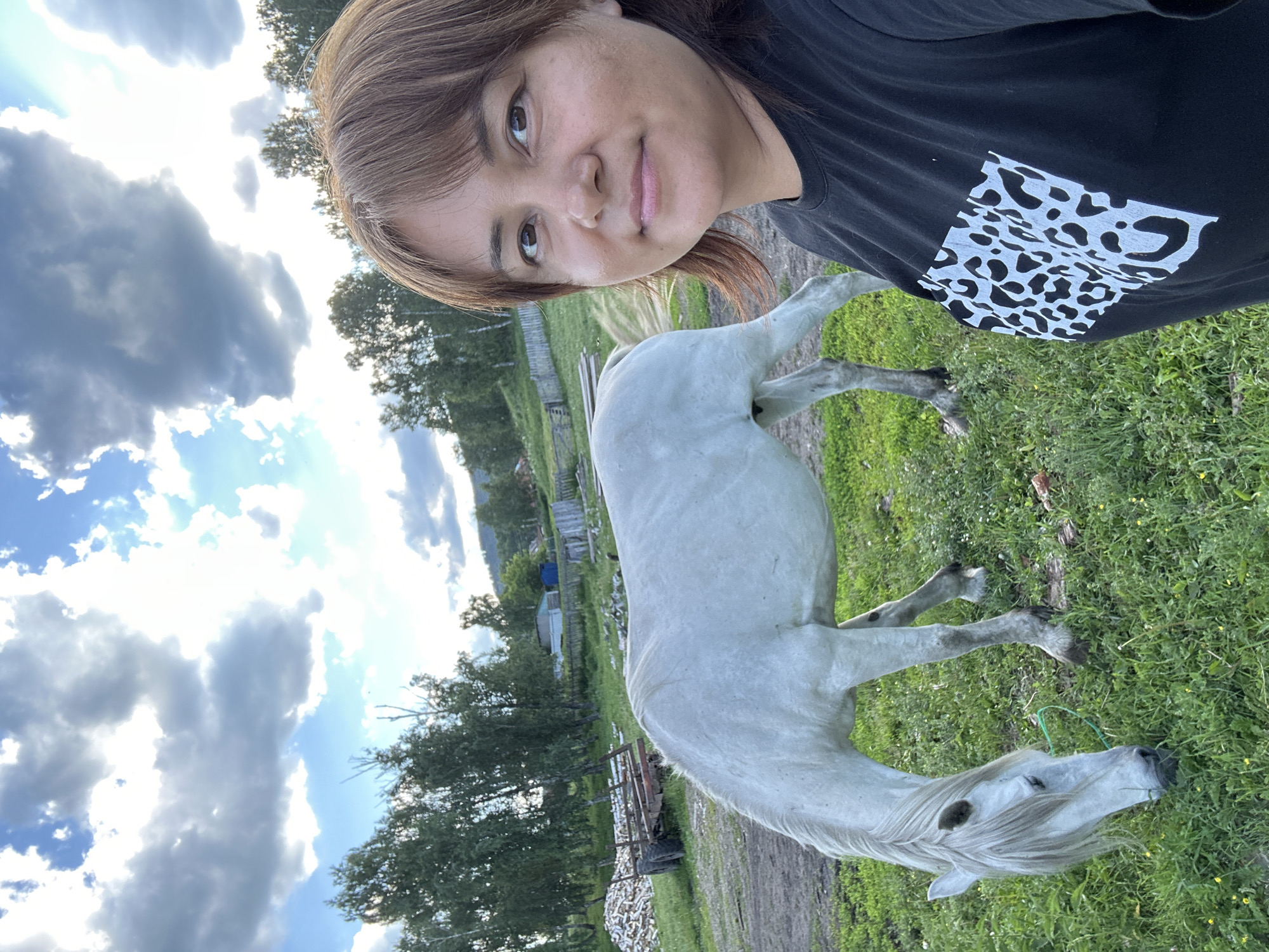 A woman is taking a selfie with a white horse in an outdoor, rural setting. The woman, with shoulder-length brown hair and bangs, smiles at the camera, wearing a black t-shirt with a white abstract pattern on the left side of the chest. She appears to be in good spirits. The horse, with a light gray coat and flowing white mane, stands to her right, facing away from the camera and towards the right side of the frame. It appears to be grazing on grass.
The scene is set in a grassy field with trees in the background. Some wooden fences and structures are also visible, suggesting a farm or stable environment. The sky is partly cloudy with bright sunshine, indicating it's likely daytime, possibly late afternoon given the lighting. The overall atmosphere is casual and relaxed. The image is oriented vertically, with the top of the frame showing the sky and the bottom showing the horse's head and the grassy ground.