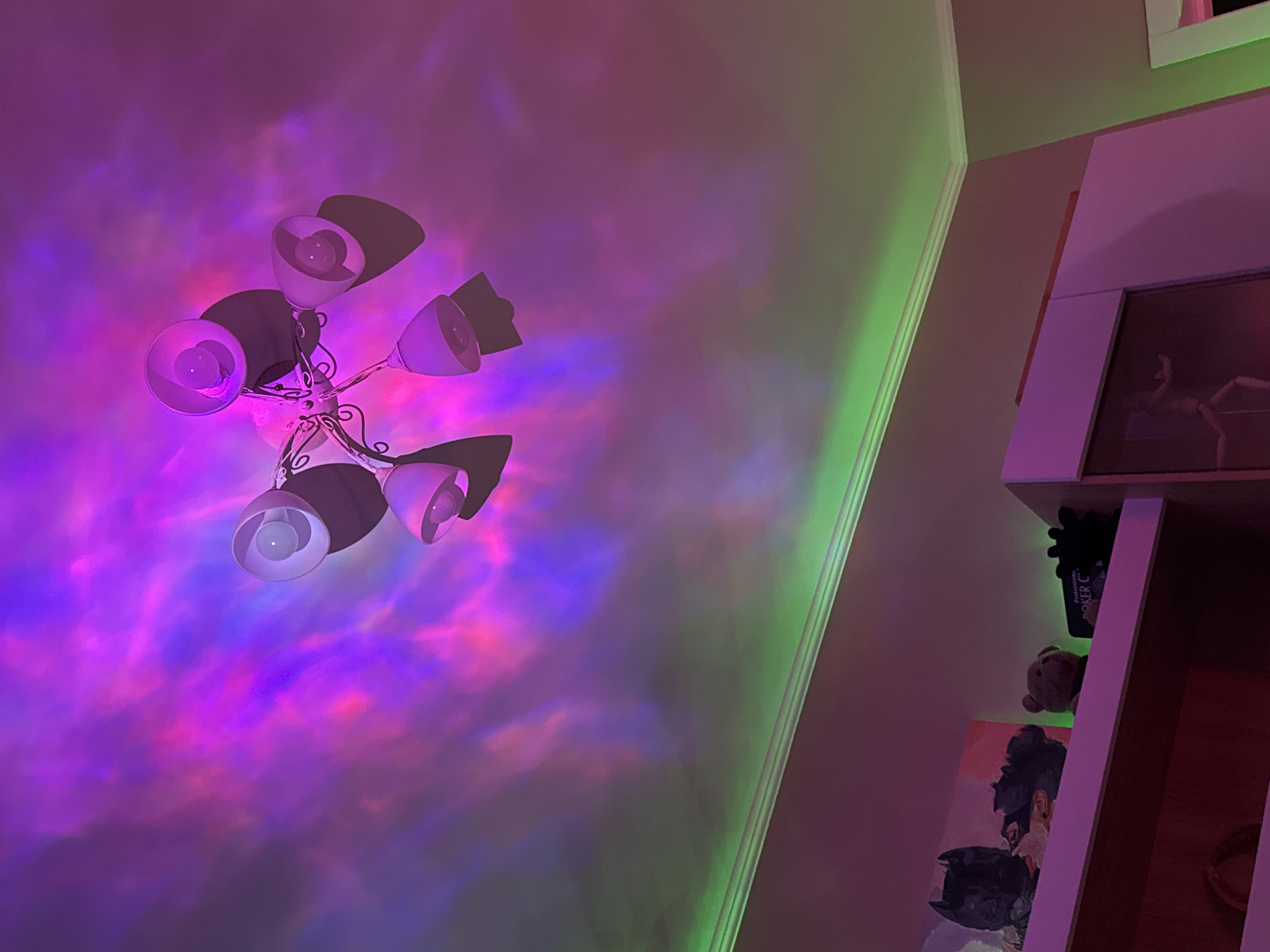 This indoor shot captures a room illuminated by dynamic, colorful lighting, creating an atmospheric and artistic ambiance. The setting appears to be a personal space, possibly a bedroom or living area.
The primary subject is a multi-bulb light fixture, likely a chandelier, mounted on the wall or ceiling in the upper left quadrant. It features several ornate, metallic arms supporting individual, cup-shaped lampshades with visible bulbs. These bulbs cast light, but the dominant illumination comes from other sources.
The wall and ceiling behind the fixture are bathed in swirling patterns of purple, pink, and blue light, resembling a nebula or ocean effect. This suggests the presence of a light projector creating these intricate, moving patterns. Along the seam where the wall meets the ceiling on the right side, a strip of vibrant green light, likely an LED strip, casts a distinct glow on the architectural trim.
On the right side of the image, a vertical white or light-colored shelving unit is visible. On its top shelf, there is a framed picture or artwork depicting a light-skinned figure in a dynamic pose. The middle shelf holds a dark, plush-like object, a small teddy bear, and a box that partially displays the text "Protection" and "POKER C". The lowest visible shelf features a painting or print of a stylized head and shoulders, with what appears to be a black bat symbol on the chest area. A small, circular object, possibly a ring or bracelet, also rests on this shelf.
No specific activity or event is taking place; the image is a still capture of the room's decorative lighting setup. The extensive use of artificial, colorful lights indicates it is likely evening or night. The lighting creates a calm, imaginative, and perhaps playful mood within the space.