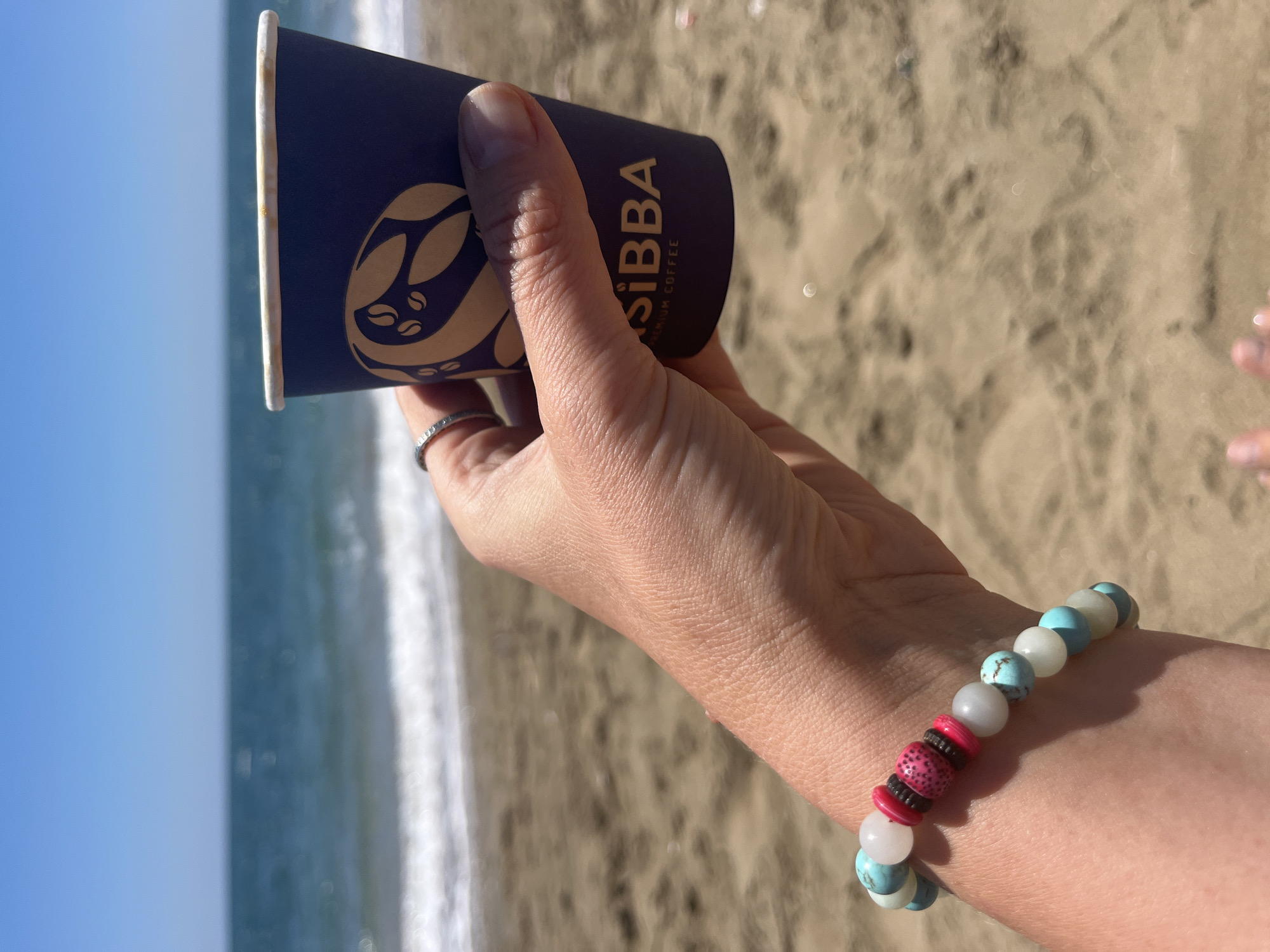 A sunlit, close-up shot features a person's hand holding a dark blue disposable coffee cup at a beach in Trikomo, Northern Cyprus. The hand, adorned with a silver-toned ring on the ring finger, also wears a beaded bracelet on the wrist. The bracelet consists of light blue and white spherical beads, with a central cluster of textured pink and dark brown disk-shaped beads.
The coffee cup displays a golden-yellow logo of stylized coffee beans and the prominent brand name "SIBBA," with "PREMIUM COFFEE" written beneath it in smaller text. The background, largely out of focus, reveals a bright blue sky, the gentle waves of the sea, and light brown sand. The strong sunlight casts distinct shadows on the hand, indicating it is daytime. The overall setting is serene, capturing a moment of enjoying a drink by the sea.