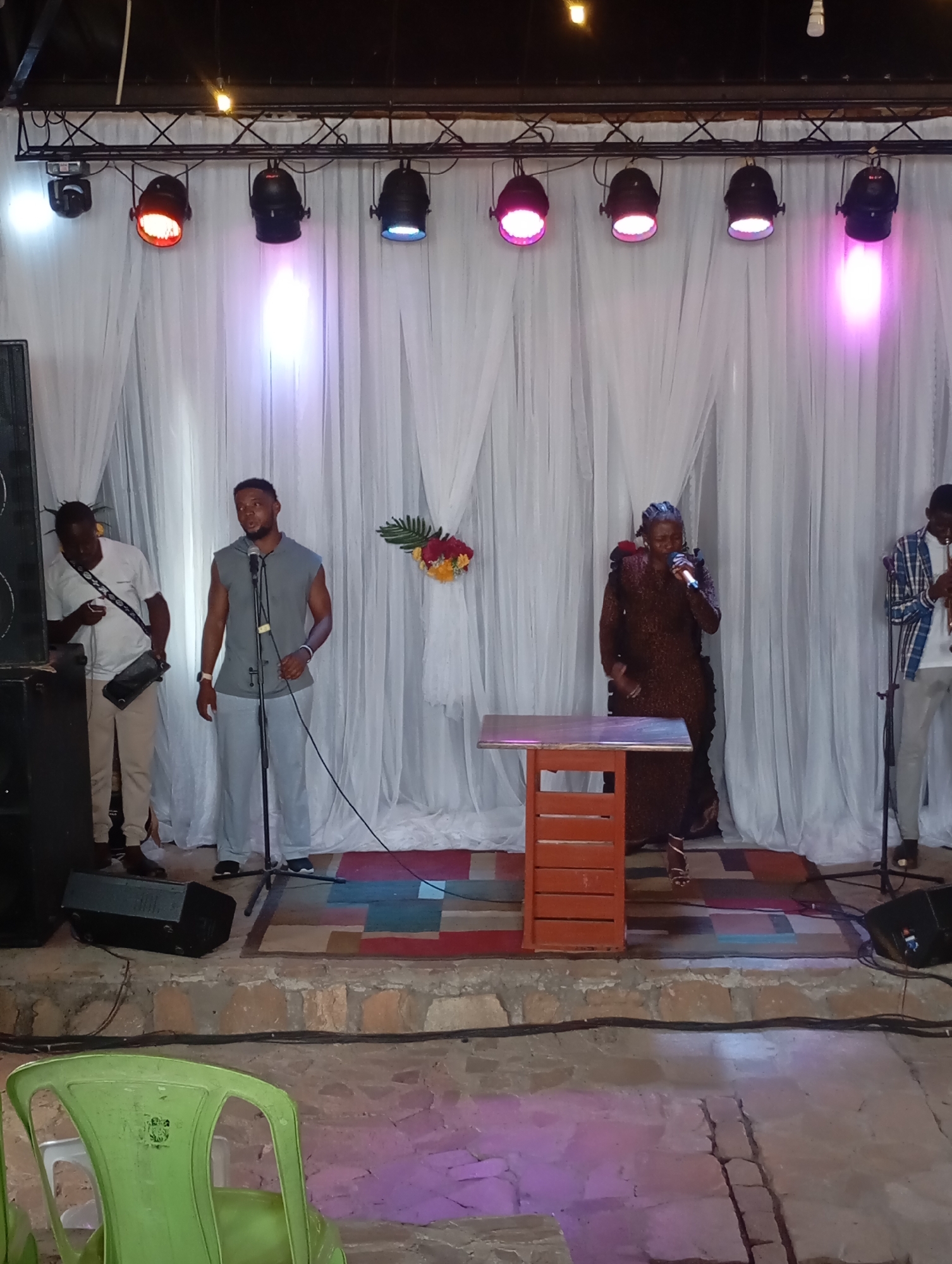 The image depicts a live performance in Uke, Nigeria. Four individuals are on a raised stage with a white curtain backdrop illuminated by a series of overhead stage lights. The lights emit different colors, including red, blue, and pink. From left to right, there is a man standing by a large speaker holding a black bag, a man at a microphone, a woman in a brown patterned dress singing into a microphone near a small wooden podium, and a man standing at a microphone holding an instrument, which is partially out of frame. A green plastic chair is visible in the foreground, positioned in front of the stage. A geometric patterned rug covers the central part of the stage floor.