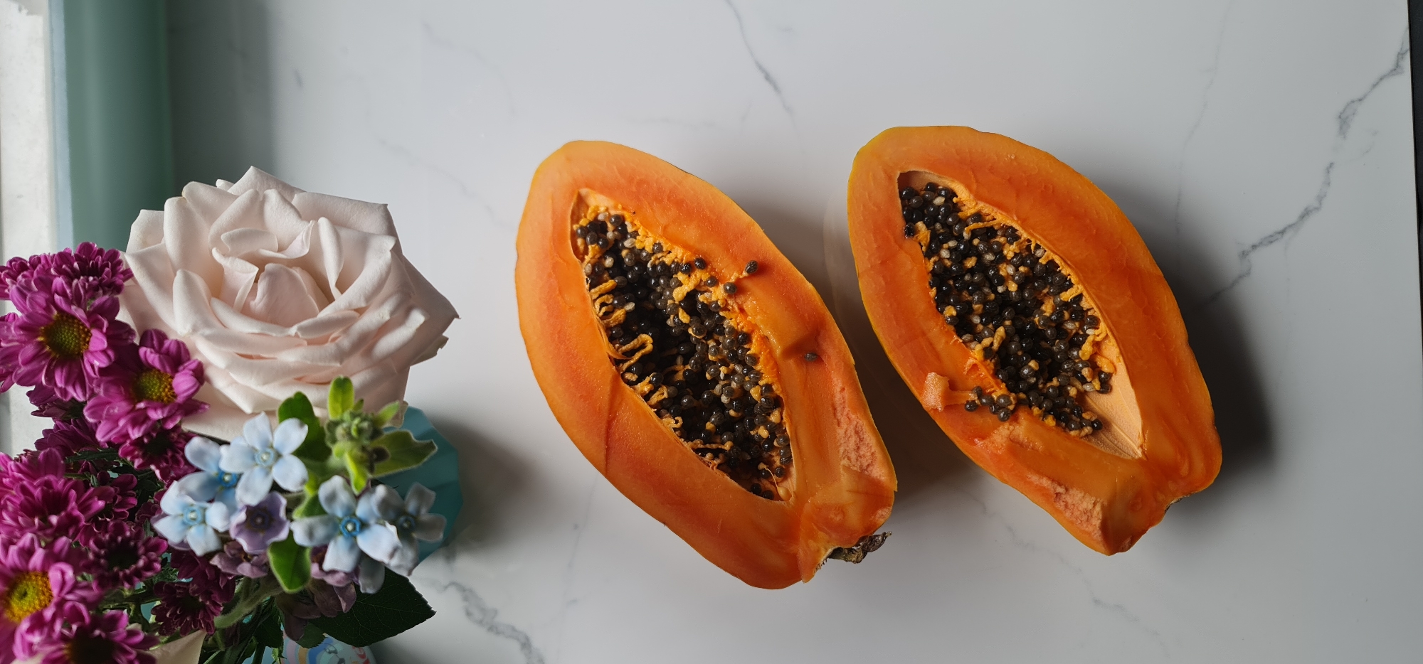 The image displays a still life arrangement on a white marble surface. Two halves of a papaya fruit, with their characteristic orange flesh and black seeds, are centrally positioned. To the left, a small bouquet of flowers is arranged in a light blue vase. The bouquet includes a large, pale rose, purple daisies, and small blue flowers. The background includes a hint of a light green element, possibly a wall or fixture. The scene is well-lit, highlighting the textures and colors of the fruit and flowers. There is no indication of the location being Đà Nẵng, Vietnam or any people, actions or interactions.