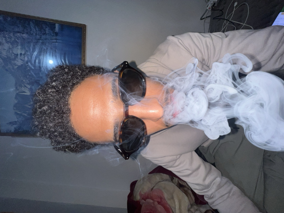 An individual with dark, curly hair and a medium complexion is depicted in a medium close-up, exhaling a dense plume of white smoke from the mouth. The person wears tortoise-shell framed sunglasses with dark lenses, obscuring the eyes. A reddish tint is visible on the lips. The subject is dressed in a light-colored, long-sleeved garment. The scene is an indoor environment. A light-colored wall forms the background, featuring a framed picture of a blue-toned landscape, possibly mountains and water, with a bright reflection, hanging to the right of the subject's head. To the left, a section of a bed or sofa covered with a reddish-brown blanket, patterned with lighter shapes, is visible. White cables are present on a dark surface to the right. The general location is within the city of Tanger, Morocco; however, the specific address or type of establishment is not identifiable from the image.