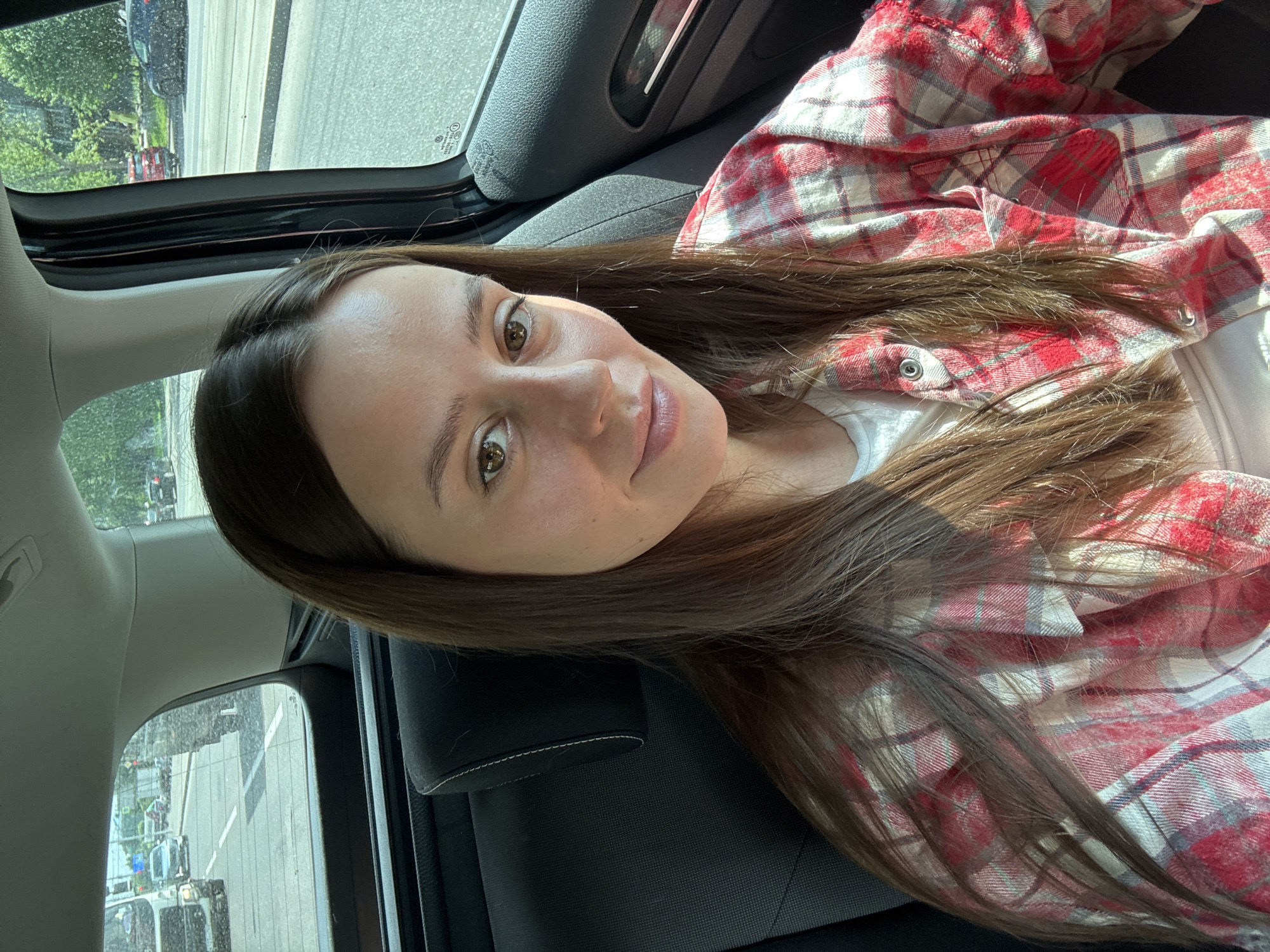 In the image, a young woman with long brown hair is seated in the back of a car, looking towards the camera. She is wearing a red and white plaid shirt over a white top. The interior of the car is visible, including a black seat and headrest. Sunlight filters through the windows, casting shadows on her face and hair. Through the car windows, a city street scene can be observed. Cars are driving on the road, and trees and buildings line the street. The overall impression is that of a candid snapshot taken during a car ride in an urban environment. The specific location is not identifiable from the image alone, however, based on common elements in such images and the likely origin of the image, it could be a street in Krasnoyarsk, Russia.