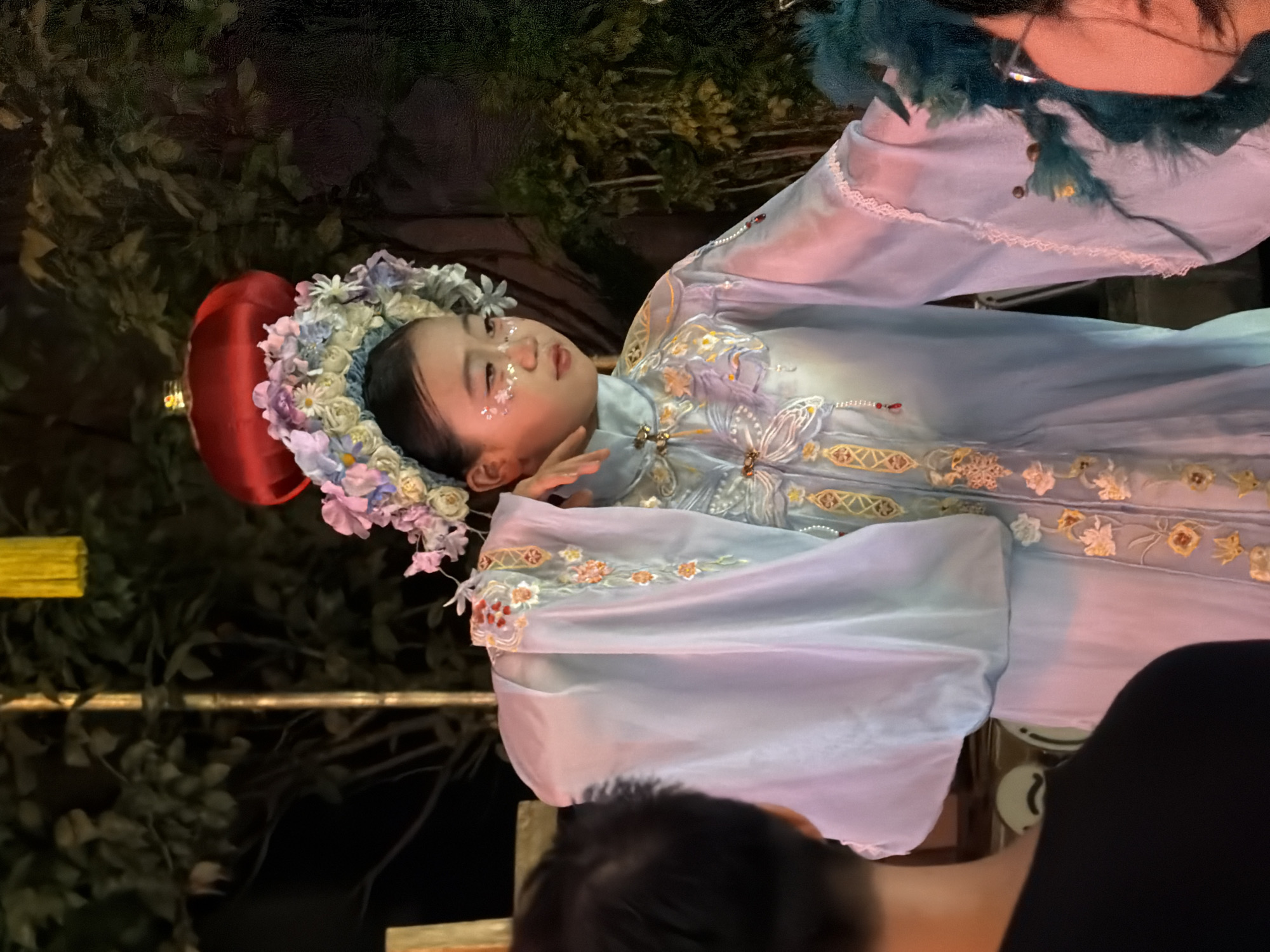 In the Yuzhong District of China, a young girl, adorned in a light blue traditional Chinese dress with intricate embroidery and floral headwear, performs. The dress features long, flowing sleeves and detailed embellishments of flowers and ornate patterns. Her face is decorated with glitter, adding a festive touch to her performance. She stands in what appears to be an outdoor setting at night, with lush green foliage forming a dark backdrop. A red lantern is visible above her, and a yellow decorative element hangs in the upper part of the frame. In the foreground, the back of a person wearing a black shirt is visible, facing away from the camera and towards the performer. To the right, another individual is partially visible, with a hand adorned with a blue feathered prop. The overall scene suggests a cultural performance or celebration.