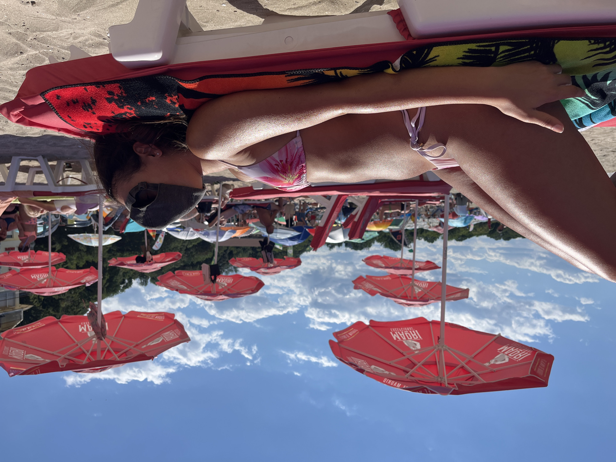 An unidentifiable person, wearing a grey baseball cap backwards, is lying on a red lounge chair on a sandy beach. They are wearing a pink bikini with a floral pattern. The person is lying on their side with their face turned away from the camera. Their arms are resting on the chair. The chair is covered with a colorful towel. In the background, there are several red umbrellas with text that reads "IRIDAM". Other beachgoers and lounge chairs are visible in the distance. The sky is blue with scattered white clouds. The location is Kiten, Bulgaria.