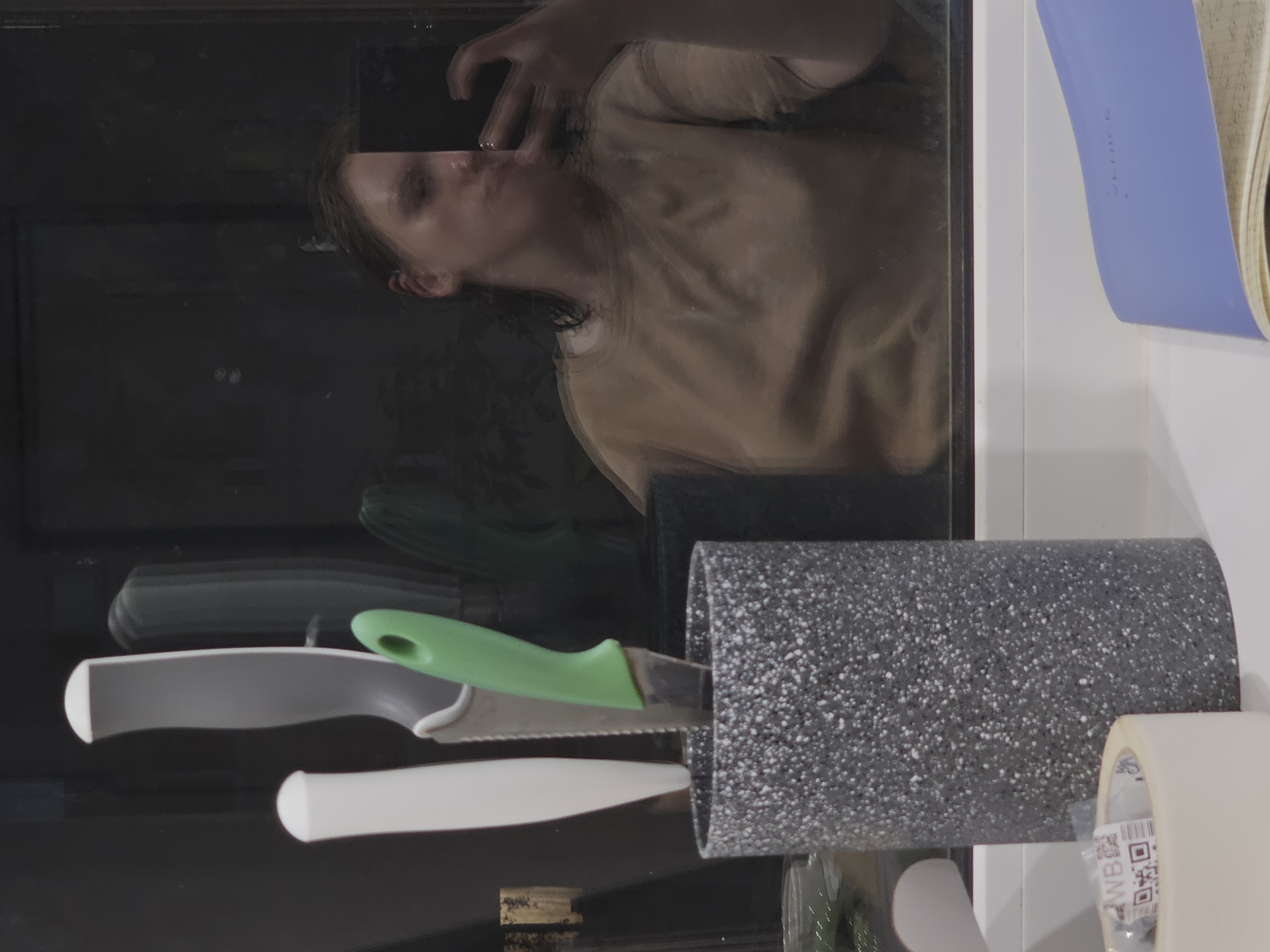 The image is a photograph taken indoors, reflecting a person holding a smartphone to take a selfie. The person is wearing a tan t-shirt. In the foreground, there is a speckled grey utensil holder with three knives in it: one with a white handle, one with a grey handle, and one with a green handle. To the left of the utensil holder, there is a roll of tape with some labeling on it, including QR codes. In the bottom right corner of the image, a portion of a light blue notebook is visible. The background is dark and appears to be a reflective surface, possibly a window or a mirror, showing the reflection of the person and the room. The environment appears to be a domestic setting, possibly a kitchen given the presence of knives. There is no visible indication of the city of Tosno, Russia in the image.