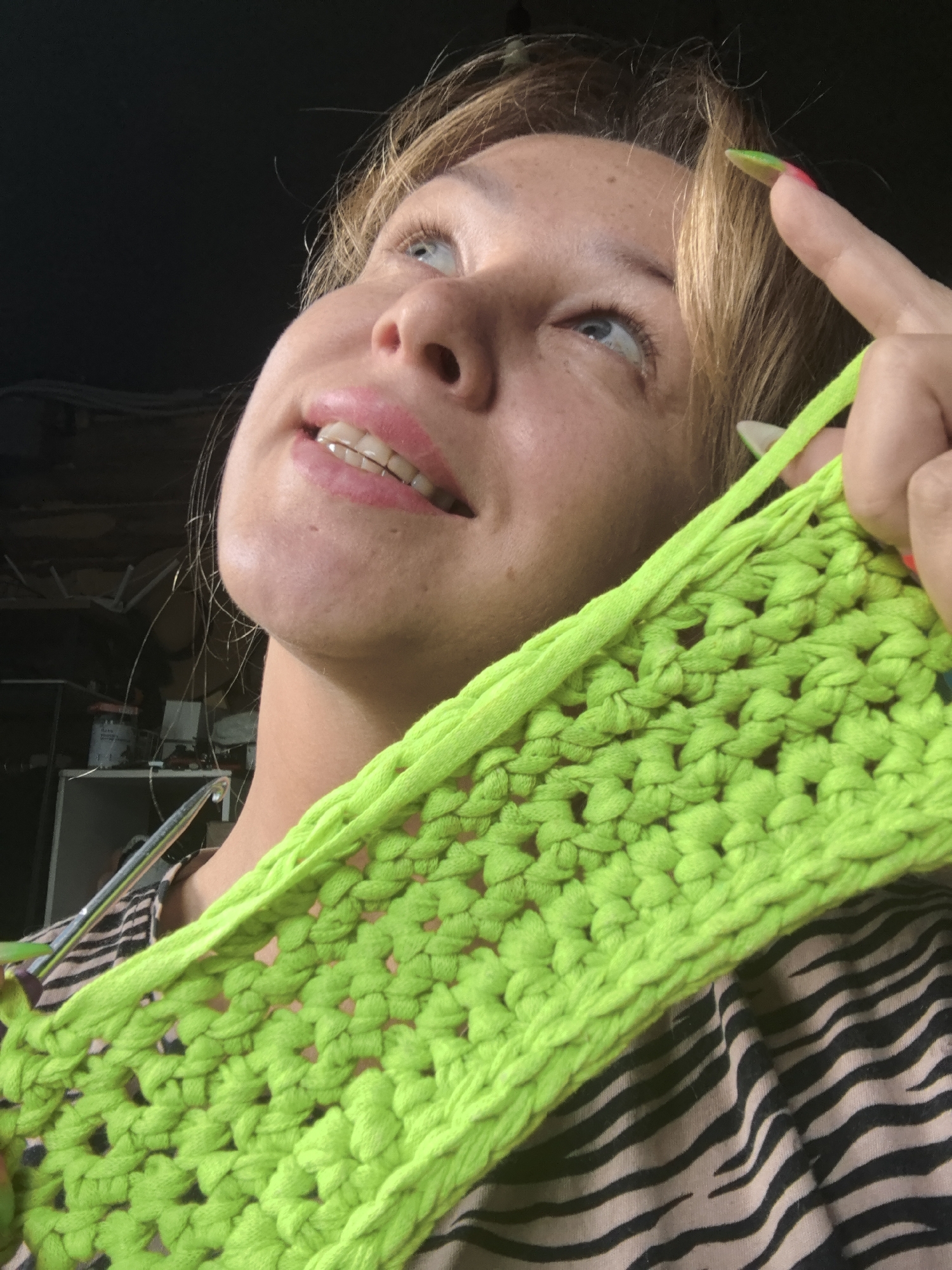 A close-up, upward-angled shot captures a woman engaged in crocheting or knitting, located in Noviy, Russia. She has fair skin, light brown hair, and bright blue eyes, which are directed upwards and slightly to the right. Her face shows a gentle smile, with her lips slightly parted, revealing teeth that appear to have clear aligners or retainers. Freckles are visible across her nose and cheeks.
She is holding a vibrant, neon green piece of chunky crocheted or knitted fabric in her left hand, which dominates the lower right side of the frame. Her fingernails exhibit a distinctive multi-colored design, blending green/yellow at the tips into pink/orange. A metallic crochet hook or knitting needle is partially visible near her left shoulder, indicating the crafting activity.
The woman is wearing a top with a black and light brown/beige zebra print pattern. The background is largely dark and out of focus, suggesting an indoor setting with strong directional lighting, possibly from a window or overhead source, highlighting her face and the bright green craft material. There is no visible text in the image.