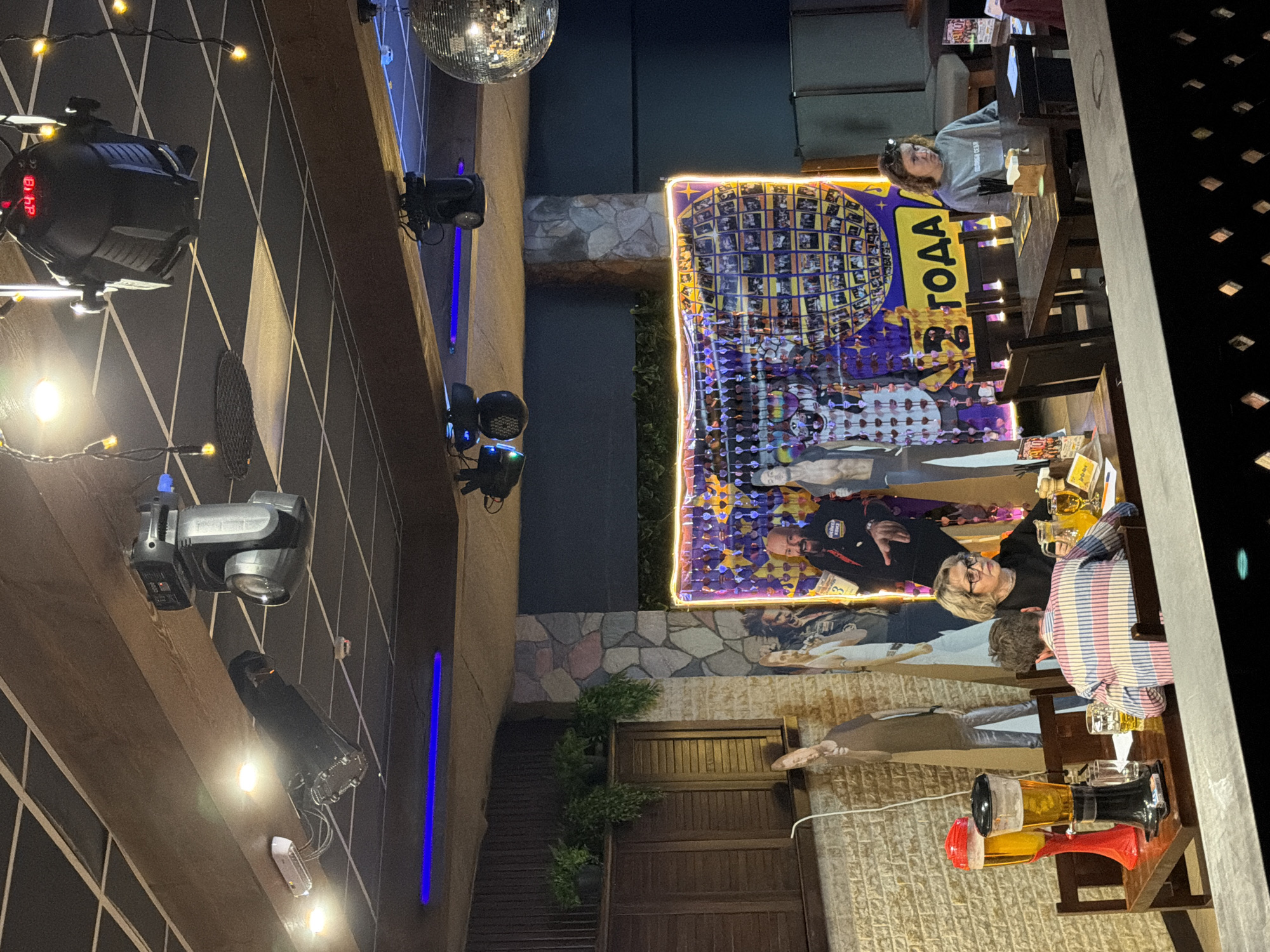 The image shows an indoor or semi-outdoor setting, possibly a bar or restaurant in Chaikovsky, Russia. The presence of a disco ball, stage lighting equipment, and a banner suggest the location is set up for an event or celebration. The banner prominently displays the word "года" (years) in Russian, possibly indicating an anniversary or commemorative event. People are seated at tables, some engaged in conversation and drinking. The environment includes wood and stone elements, suggesting rustic or themed interior design. The scene seems to depict a social gathering in a public or semi-public venue.