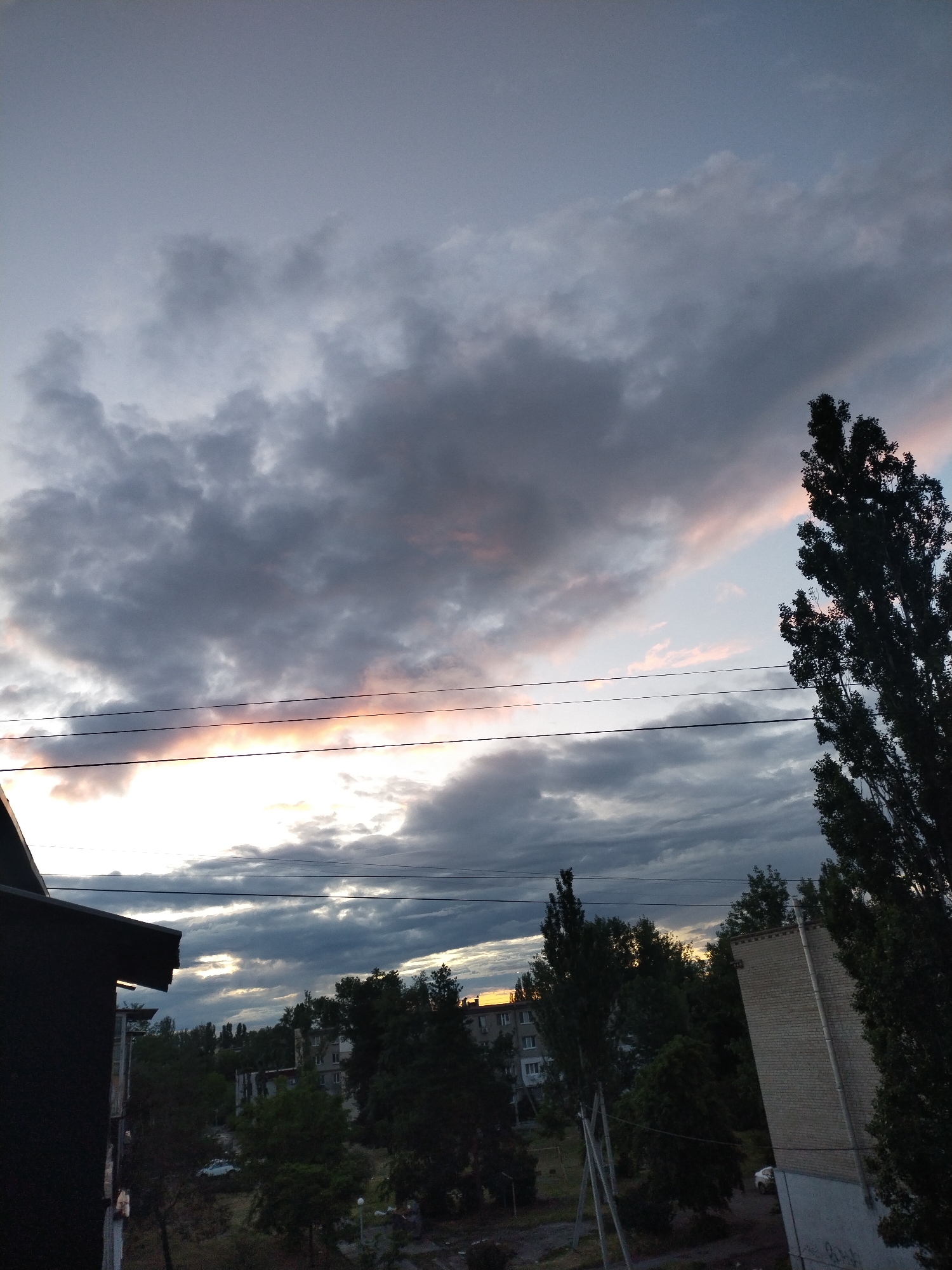 The image shows an urban landscape, likely taken from an elevated position, possibly a balcony or higher floor of a building, in Horishni Plavni, Ukraine. The sky is dominated by dramatic cloud formations, with colors ranging from grey to pinkish-orange near the horizon, suggesting either sunrise or sunset. Several power lines stretch across the sky. Below, trees and buildings are visible, creating a silhouette against the lighter parts of the sky. A brick building and the dark silhouette of another building are in the immediate foreground. The overall scene conveys a tranquil, somewhat subdued atmosphere.
