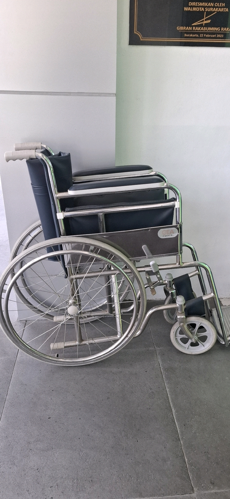 A wheelchair is parked on a tiled floor next to a white wall. A framed plaque hangs on a mint green wall to the right of the wheelchair. The plaque has black text and a gold border, and it is likely a dedication or official announcement, with the location "Surakarta, 22 Februari 2023" visible. The wheelchair is made of chrome and black fabric, with large spoked wheels and smaller front wheels. The image is taken from a slightly low angle, focusing on the wheelchair.