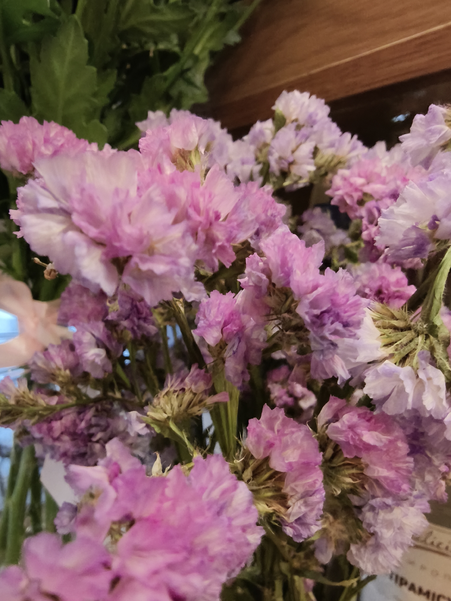 The image shows a close-up of a bouquet of pink and lavender-colored flowers. The flowers appear to be of the Limonium (Statice) type, characterized by their papery texture. Green leaves are partially visible at the top left of the image. In the lower right corner, a portion of a label or sticker with text, potentially "PARAMIC," is seen. The overall setting suggests an indoor environment, perhaps a florist shop or a home. The image contains no details of a location or the city of Kryvyi Rih.
