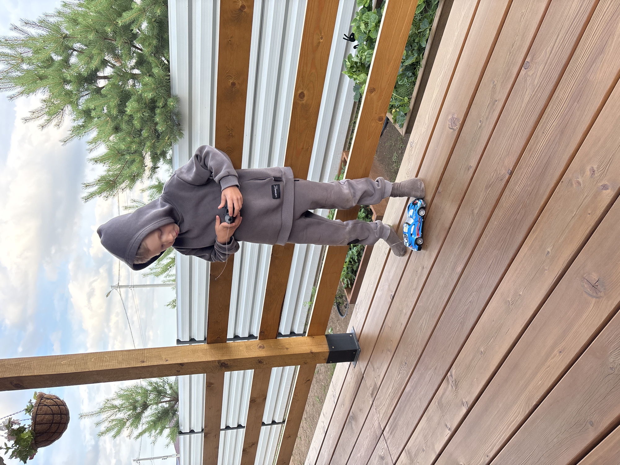 A young boy, estimated to be between 5 and 7 years old, is standing on a wooden deck, holding a black remote control in his hands. He is wearing a gray hooded sweatshirt and matching sweatpants, with gray socks. In front of him on the deck is a blue toy car. The deck is made of light brown wooden planks. Behind the deck, there is a wooden fence with horizontal planks and a corrugated metal fence. To the left of the boy, a wooden support post is visible, and hanging above it is a woven basket with flowers. Beyond the fences, there are green trees and a cloudy sky. The scene appears to be an outdoor residential area.