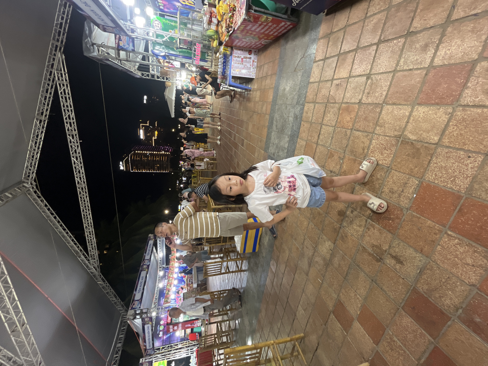 The image depicts an outdoor night market scene in the city of Đà Nẵng, Vietnam. In the foreground, a young Asian girl stands facing the camera, wearing a white t-shirt and denim shorts, holding a plastic bag. She is wearing light-colored sandals. Behind her, a man in a striped t-shirt and shorts is on his phone. The market features numerous stalls with food and merchandise, illuminated by bright lights. Several tables and chairs are visible, suggesting a dining area. In the background, city buildings, including a tall hotel with illuminated windows, are visible against the night sky. The ground is paved with terracotta tiles. The overall atmosphere is that of a bustling night market.