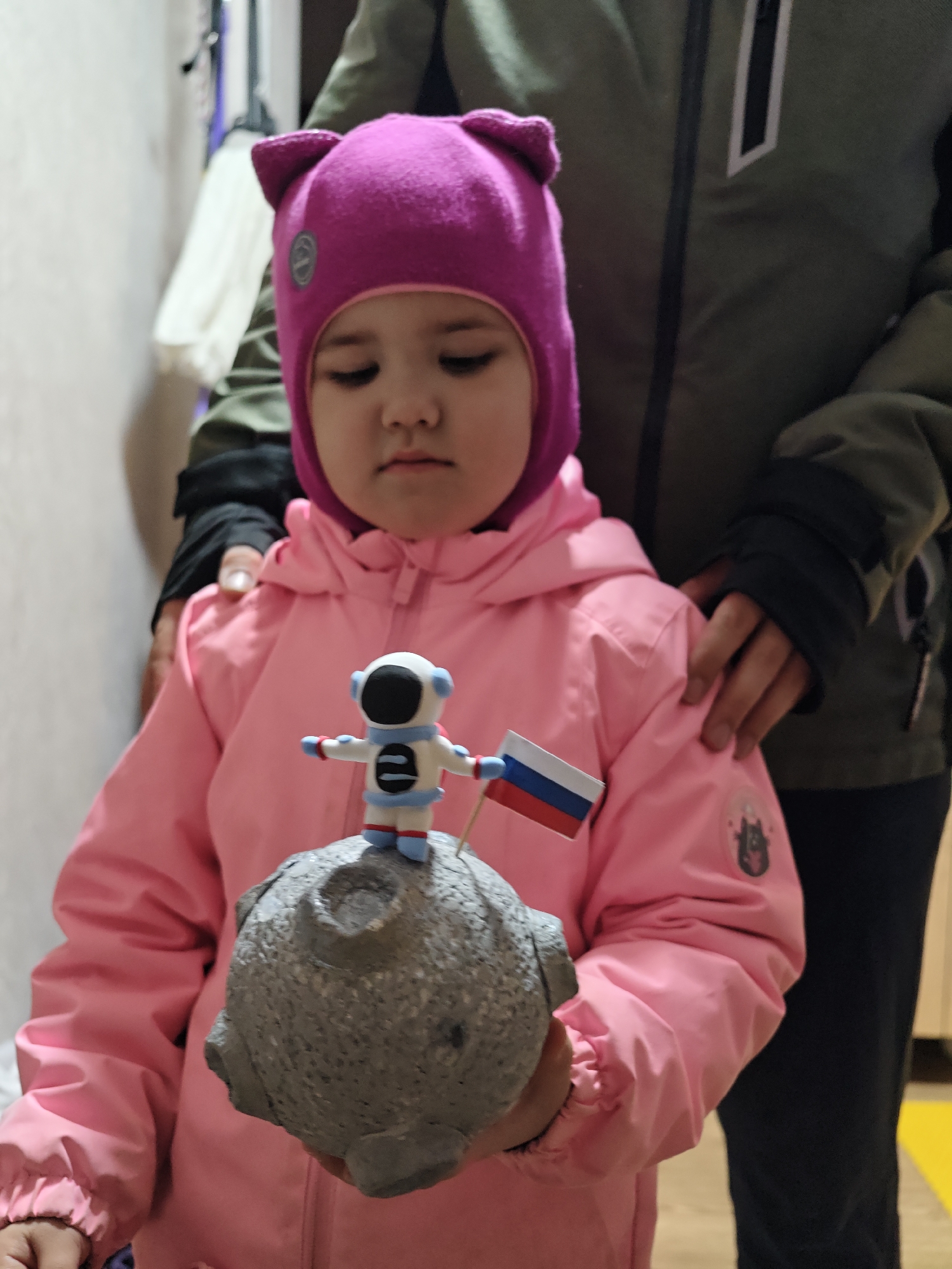 A young child wearing a pink jacket and a purple hat with cat ears is holding a gray, spherical model of the moon with a small astronaut figure standing on top and holding a Russian flag. The child appears to be indoors, possibly in a hallway. An adult is standing behind the child with their hands resting on the child's shoulders. The adult is wearing a green jacket. The location is Chaikovsky, Russia.