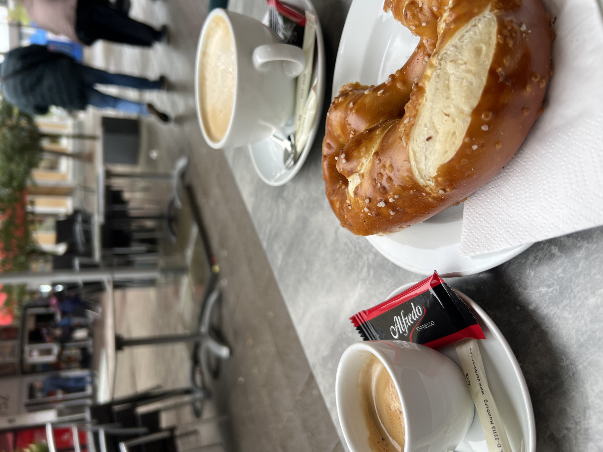 The image captures a scene at a cafe in Kehl, Germany. A pretzel rests on a white plate with a napkin. Beside it are two cups of coffee, each with a saucer and a small package of Alfredo Espresso coffee and a sugar stick. The table is outdoors, with a grey surface. In the background, blurred figures of pedestrians are visible on a sidewalk, along with buildings and trees characteristic of an urban environment. The day appears overcast, with soft, diffused light.