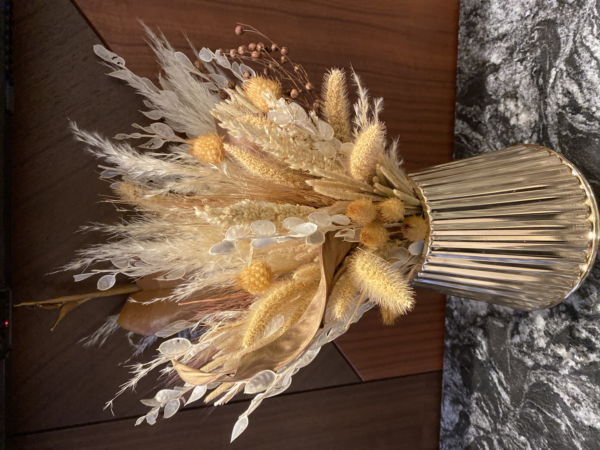 The image shows a bouquet of dried flowers in a gold vase, placed on a brown wooden surface and next to a black and white marble-like surface. The bouquet includes elements like pampas grass, lunaria, and spherical dried flowers. The ribbed gold vase is partly illuminated, creating a shiny effect. The photograph may be taken indoors, possibly in Alanya, Turkey, though there is no explicit location detail visible.