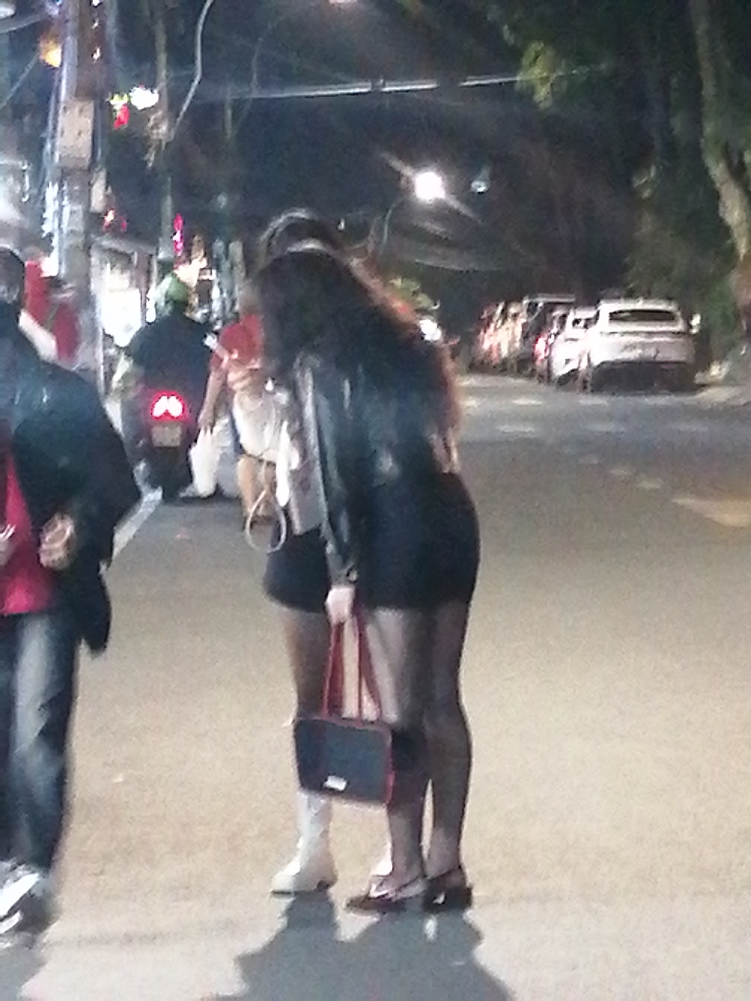 The image depicts a street scene at night in Da Lat, Vietnam. In the foreground, two individuals are partially visible. The person on the left is wearing a dark jacket and jeans. The person on the right, facing away from the camera, is wearing a black leather jacket, a dark dress or skirt, and sheer black hosiery. They are holding a dark-colored handbag with red trim. Another person, possibly wearing a white top and shorts, stands between them and a parked motorcycle. In the background, several cars are visible parked along the street, with one white car appearing to be a sedan. The street is paved, and overhead lights illuminate the scene. Trees line the right side of the street, their foliage dark against the night sky. Lights from buildings and streetlights create a diffused glow, suggesting an urban environment at night.