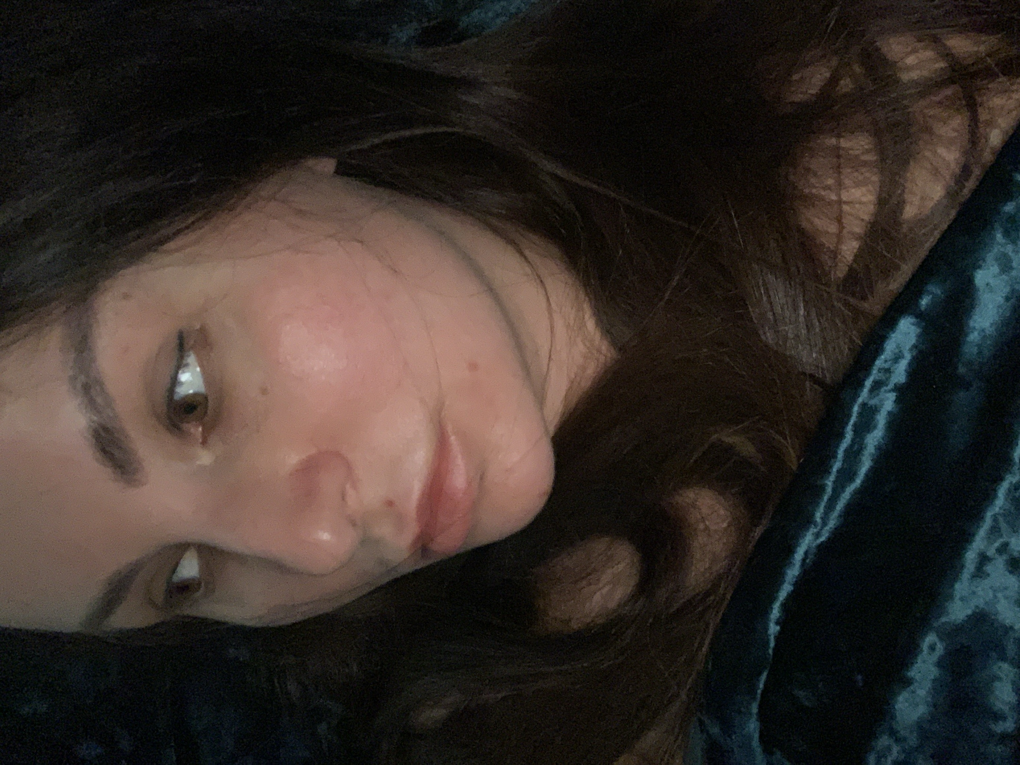 This is a close-up, low-angle, indoor shot of a person's face. The subject is lying down, with their head tilted to the left, and their eyes are looking downwards and to the left. Their dark brown hair is spread out around their face, and they are nestled against a dark teal, possibly velvet, textured fabric. The lighting is dim, suggesting it might be nighttime or a room with low ambient light. The subject has fair skin with some redness on their cheeks and a hint of blush on their lips. They are wearing eyeliner, which accentuates their eyes. The overall mood of the image is intimate and serene, possibly capturing a moment of rest or contemplation. There is no visible text in the image.