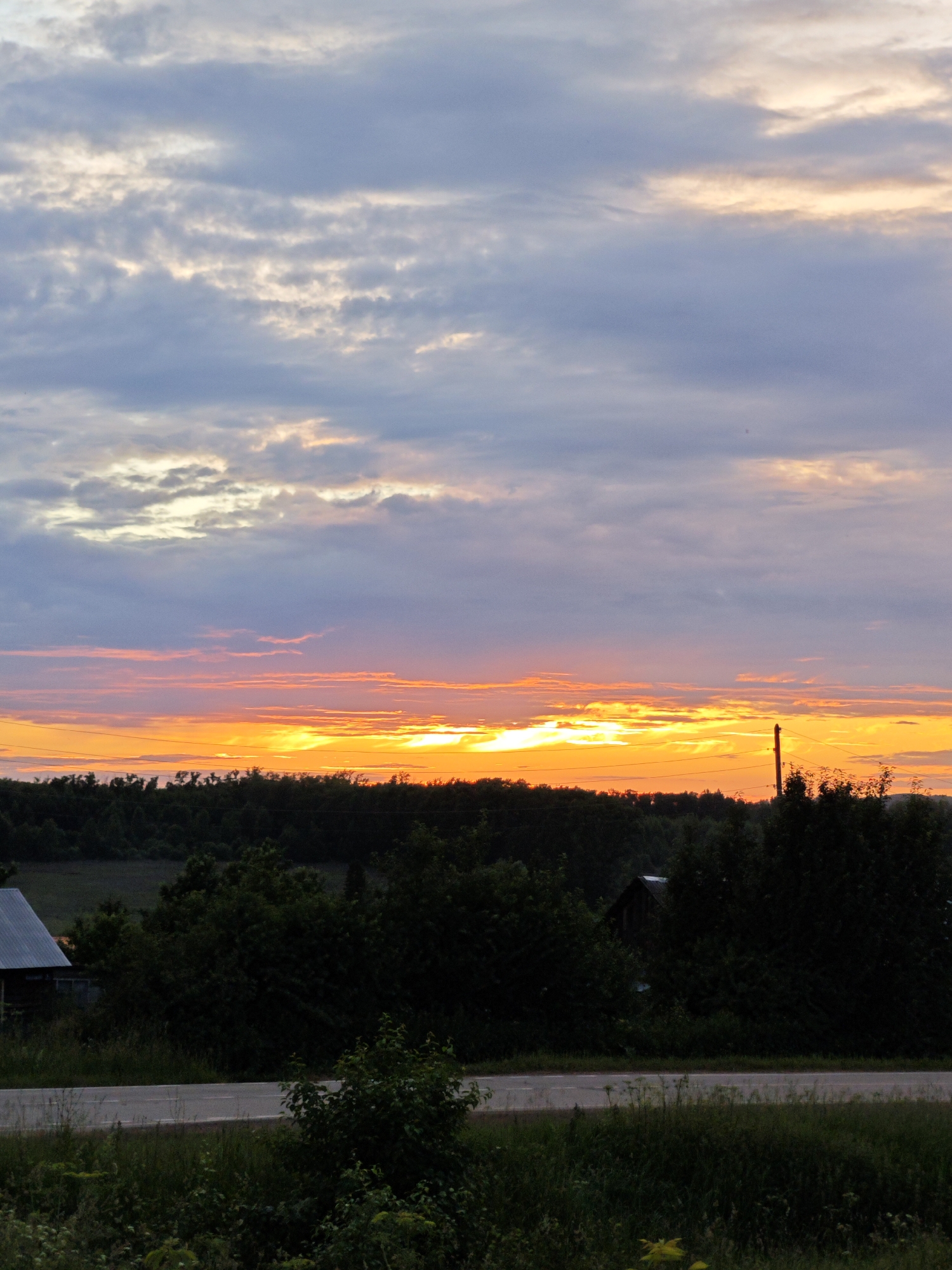 This is a vertical, eye-level, full shot of a rural landscape at sunset. The sky is filled with dramatic clouds. The bottom portion of the image is dominated by a dark, silhouetted line of trees. Below the trees, a road runs horizontally across the scene. On the left side of the road, a small, dark building with a gray roof is visible. The overall scene evokes a sense of quiet and natural beauty. The location is stated to be Foki, Russia, though no specific landmarks confirm this.