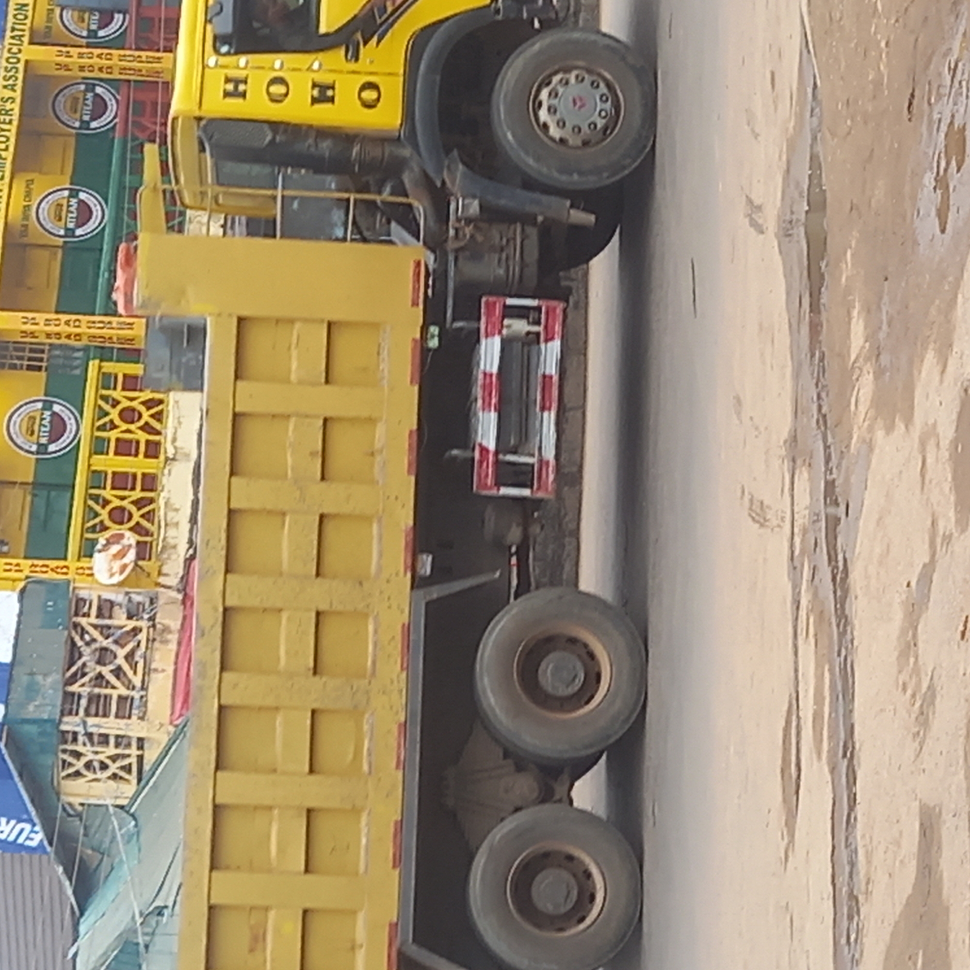 The image shows a yellow dump truck on a road in Dadi Riba, Nigeria. The truck has the word "HOHO" written on its side. It is carrying materials or goods with the names "RTLAN" and "ROAD RUPEE" on them. The background is blurred, but there are indications of buildings and infrastructure. The road surface is asphalt. The view of the truck is taken from the side, with its front facing towards the right side of the frame.