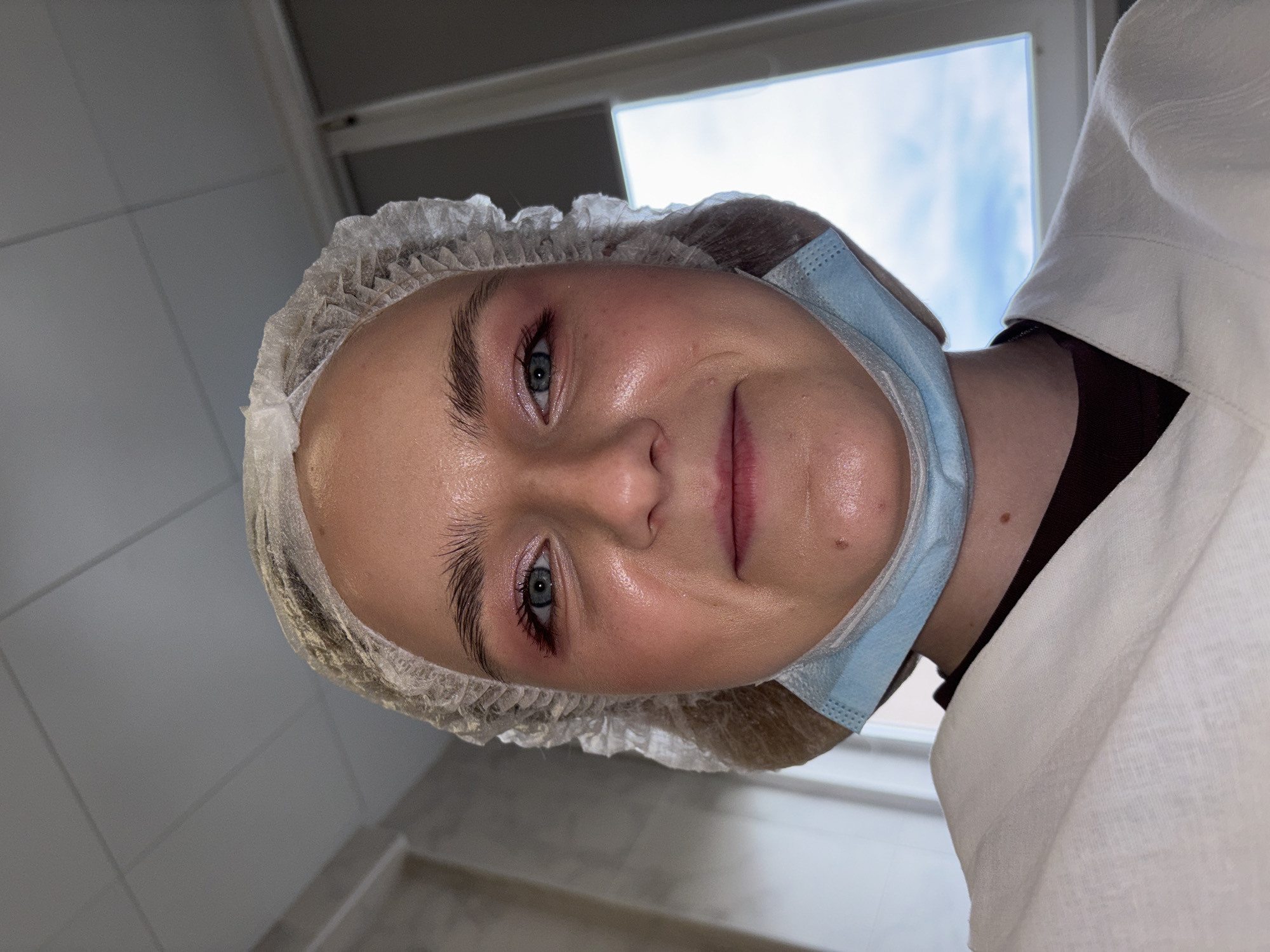The media features a young woman, positioned vertically, looking directly at the camera with a calm, neutral expression and a slight hint of a smile. She wears a white disposable bouffant cap and a light blue surgical mask pulled down below her chin. Her eyes are a light blue-grey, accentuated by mascara and subtle eyeshadow, and her eyebrows are neatly groomed. Her skin appears somewhat reflective, suggesting moisture or glow. She is dressed in a white, textured garment, likely a uniform or lab coat, over a dark undershirt.
The setting is an indoor, utilitarian space with light-colored walls and ceiling. A window in the upper right background reveals a cloudy sky, indicating it is daytime. The attire and environment suggest a professional, hygiene-conscious location, such as a medical, laboratory, or cosmetology facility. The woman appears to be posing for the photo. No visible text is present in the image. The provided context indicates the location is Saint Petersburg, Russia.