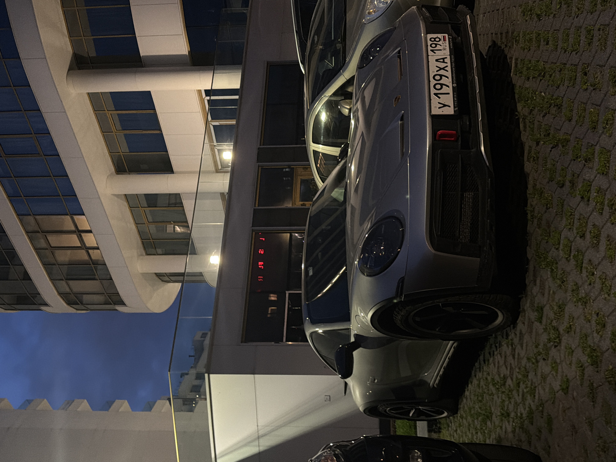 A silver Porsche 911 Carrera 4 GTS is parked on a paved surface with grass growing between the pavers, in front of a modern building at dusk. The car has black wheels and a license plate reading "Y 199XA 198 RUS". The building features large windows and white columns. Another car, a silver sedan, is parked next to the Porsche. The sky is a deep blue, indicating twilight. The scene appears to be an urban environment, likely a parking lot or street.