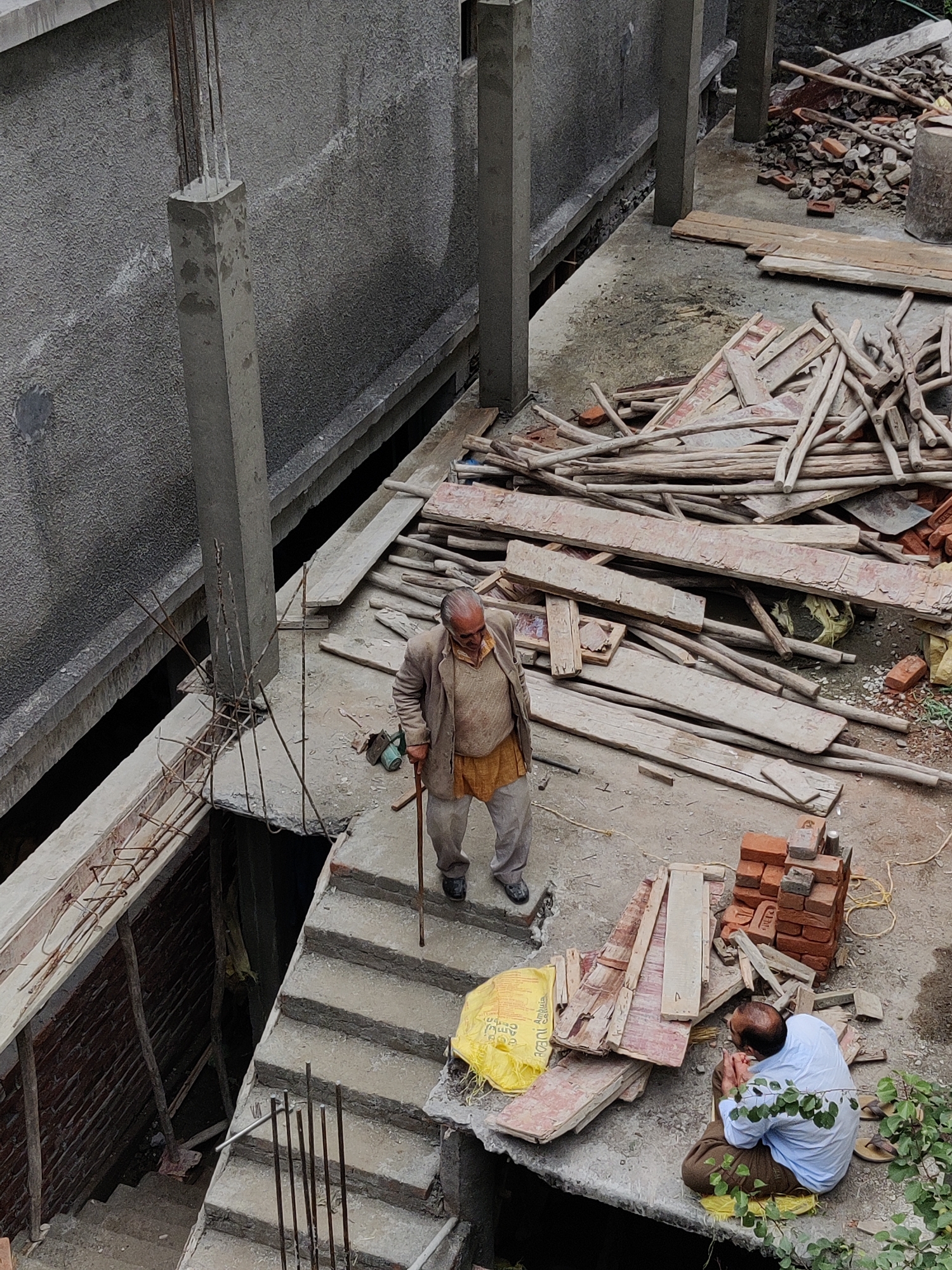 An overhead shot shows a construction site in Vashisht, India. Two men are present. In the center, an elderly man with a cane, wearing a beige jacket and orange tunic, walks down concrete stairs. To the right, a man wearing a light blue shirt and brown trousers sits on a yellow sheet, facing a pile of bricks and wooden planks. The environment is cluttered with construction materials, including lumber, bricks, and scaffolding. There is a visible concrete wall, pillars, and rebar reinforcing a concrete structure.